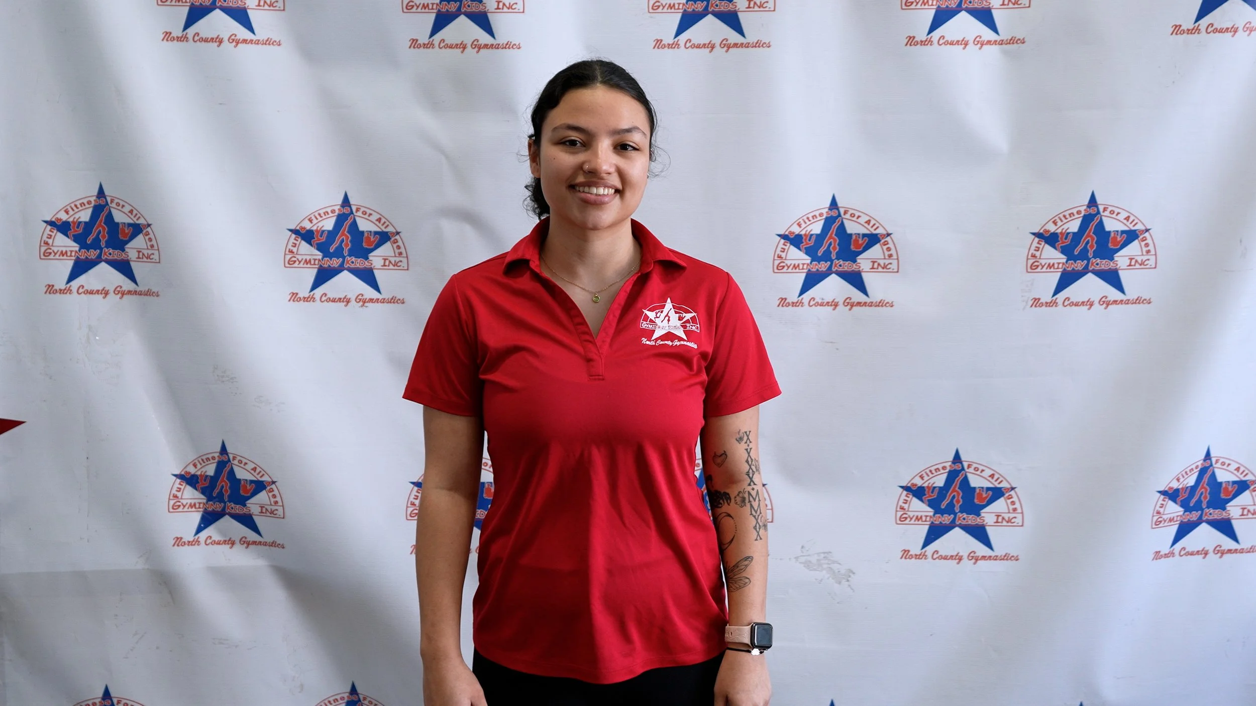 A young woman in a red shirt with the North County Gymnastics logo standing in front of a gymnastics-themed backdrop.
