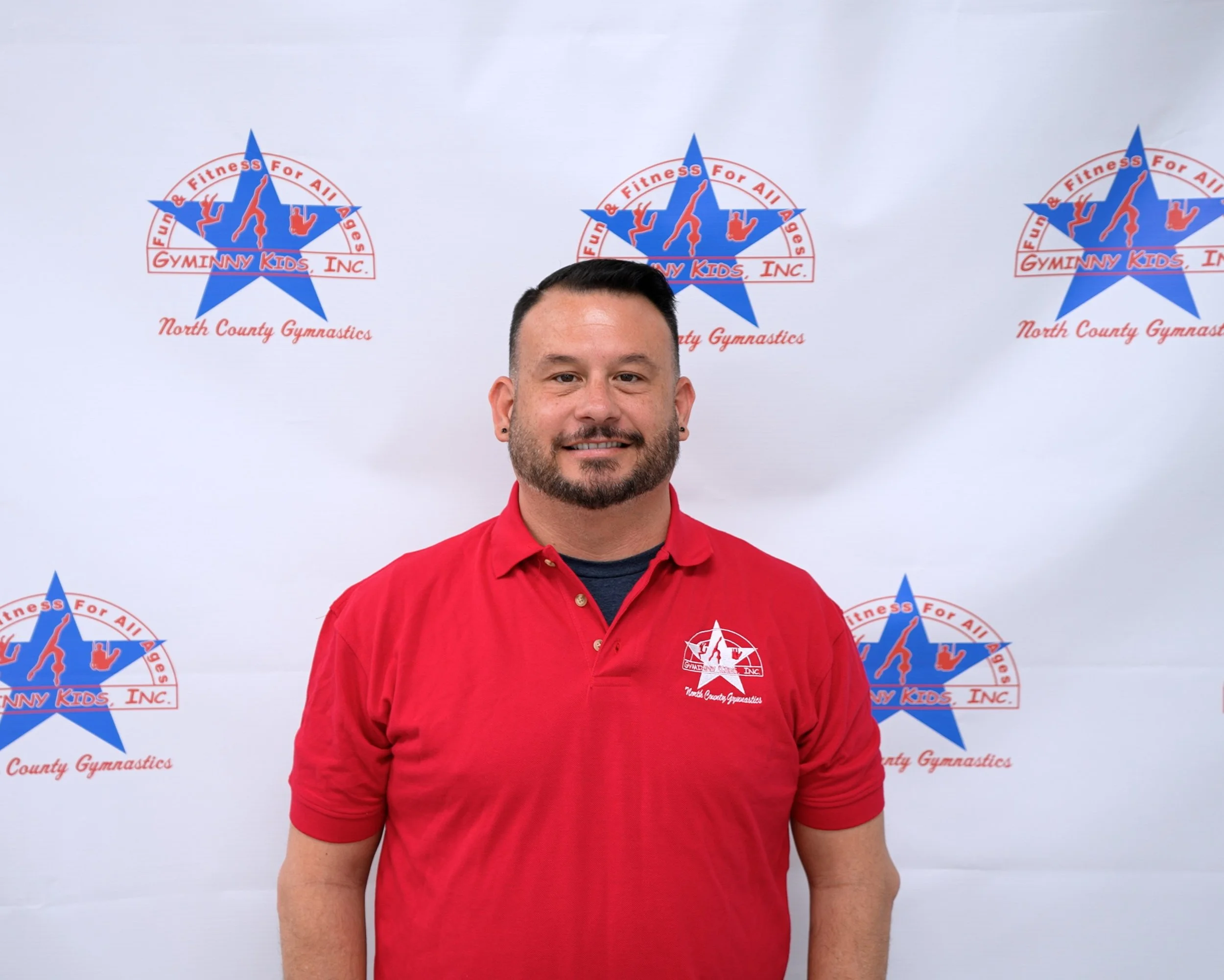 Man smiling, wearing a red polo shirt with a logo for North County Gymnastics, standing in front of a backdrop with the Gymnny Kids Inc. logo and text.