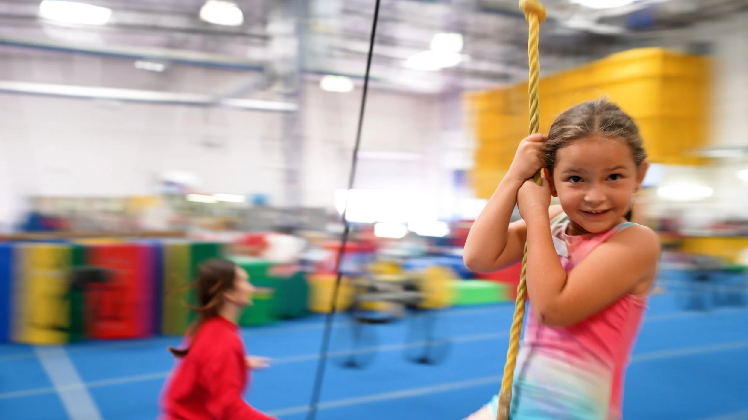 Young girl with brown hair and a pink tie-dye shirt smiling while holding onto a rope in an indoor gymnastics or trampoline park, with colorful padded walls and a blue padded floor in the background.