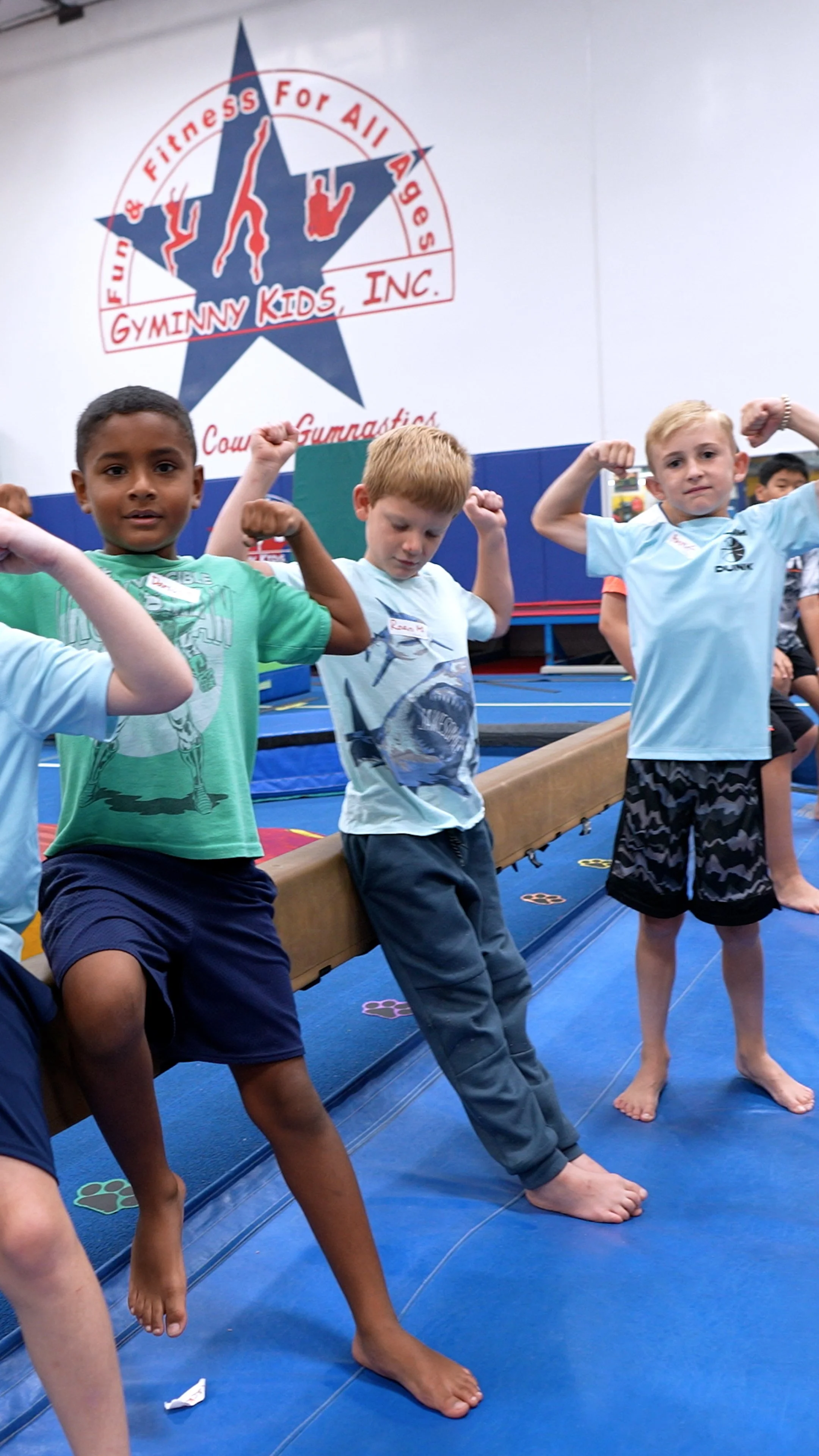 Young children at Gymminy Kids Inc. gym, flexing their arms in front of a large logo on the wall that reads 'Fun & Fitness For All Ages, Gymminy Kids Inc., Court Gymnastics.'