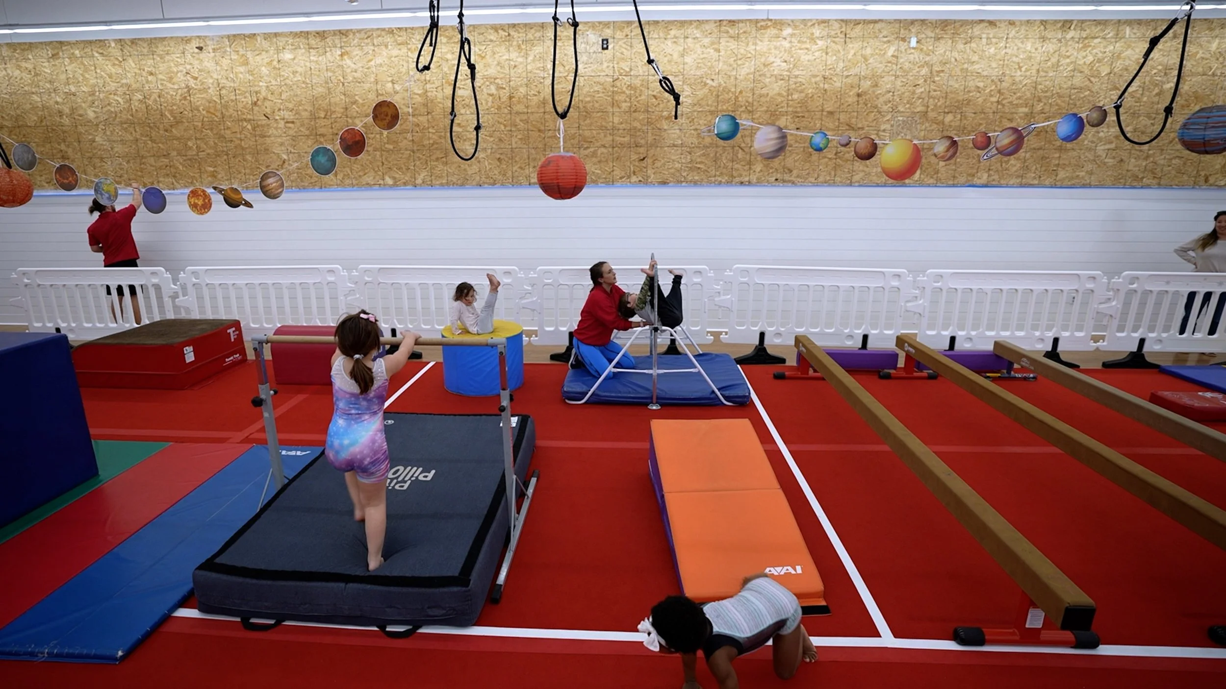 Children and adults practicing gymnastics in an indoor gym with space-themed decorations, including planets and stars hanging from the ceiling.