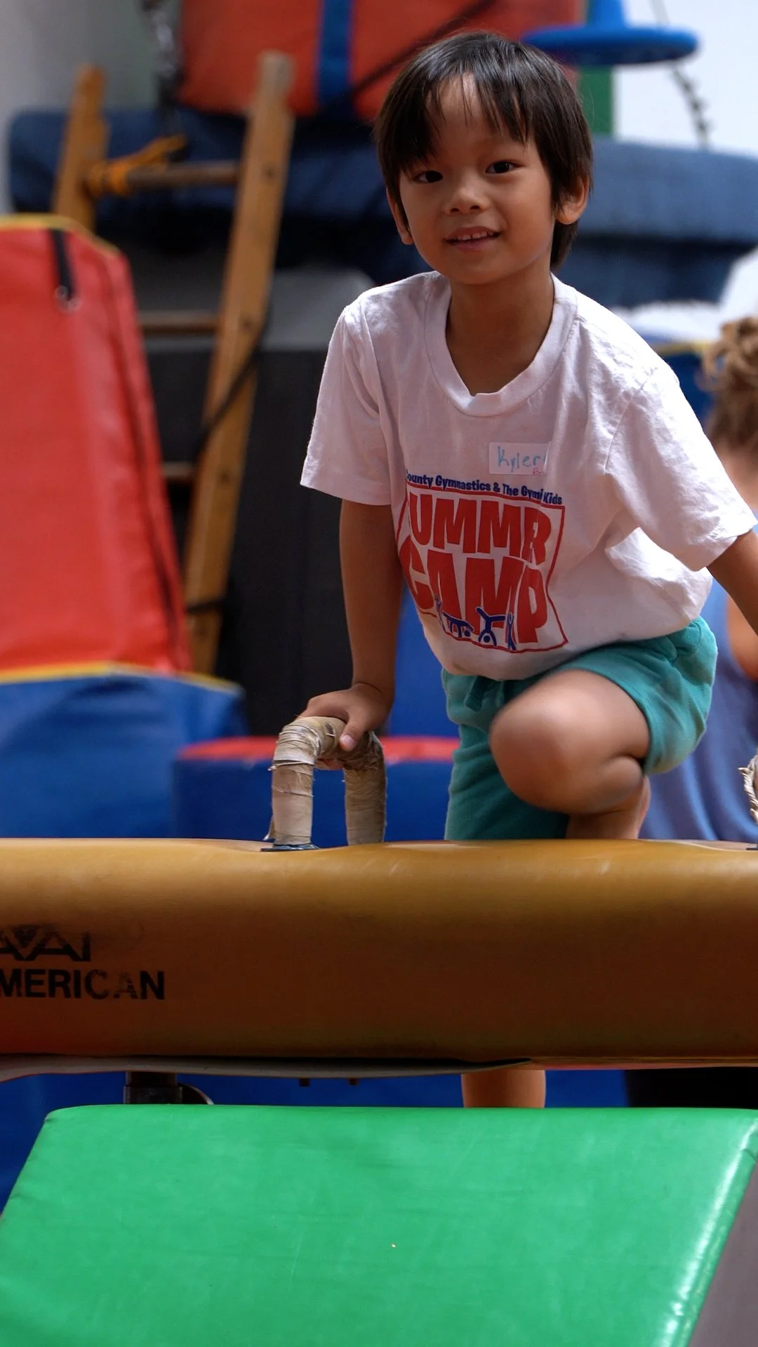 Young boy with a name tag that says Kyler, wearing a white T-shirt and teal shorts, is climbing onto an orange mat at a gymnastics summer camp.