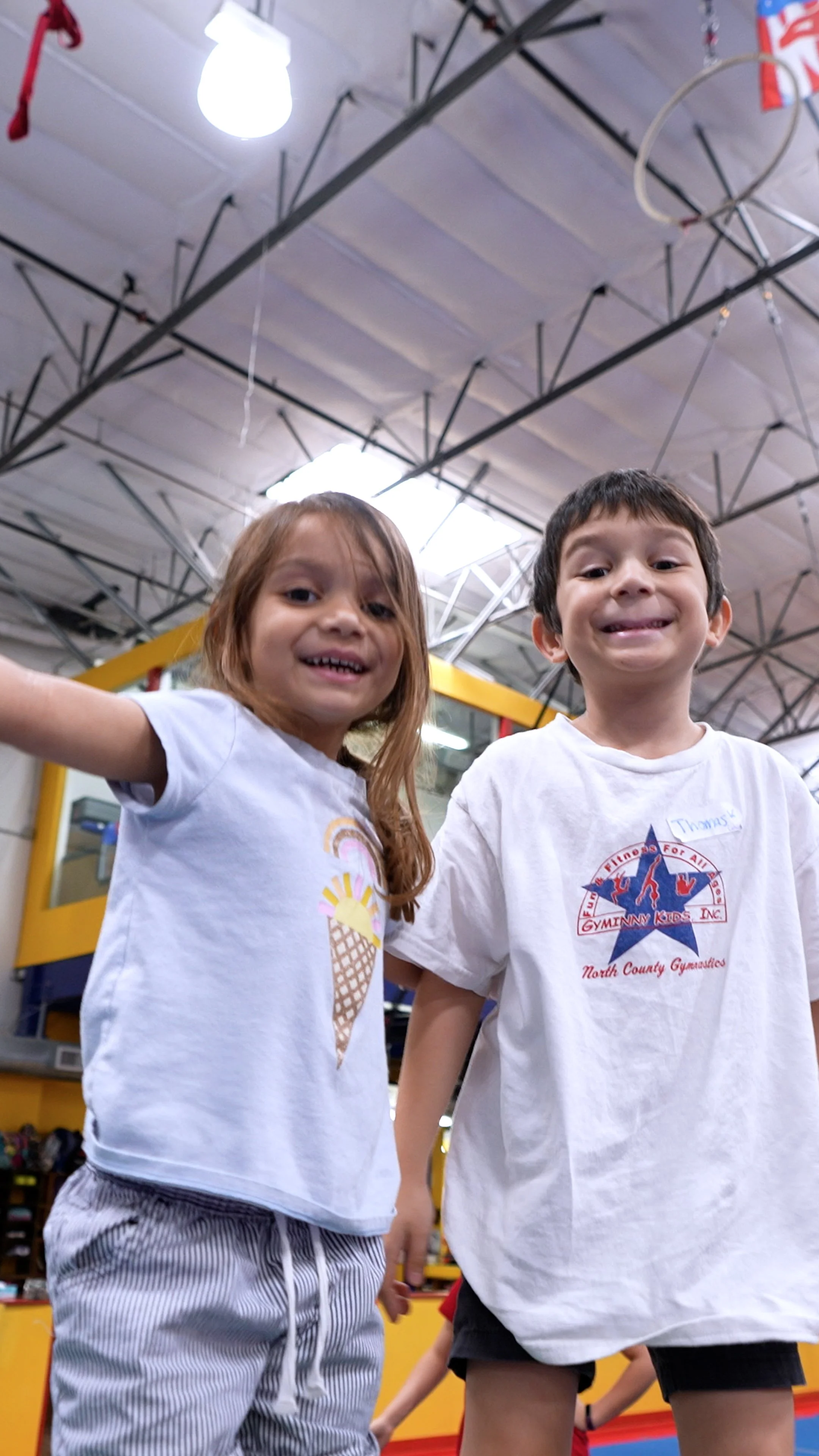 Two children, a girl and a boy, are smiling and looking at the camera inside a gymnasium or indoor play area. The girl has long, light brown hair and is wearing a white t-shirt with an ice cream cone graphic. The boy has short dark hair, wearing a white t-shirt with a red, white, and blue logo and black shorts. Both children look happy and are standing close together.
