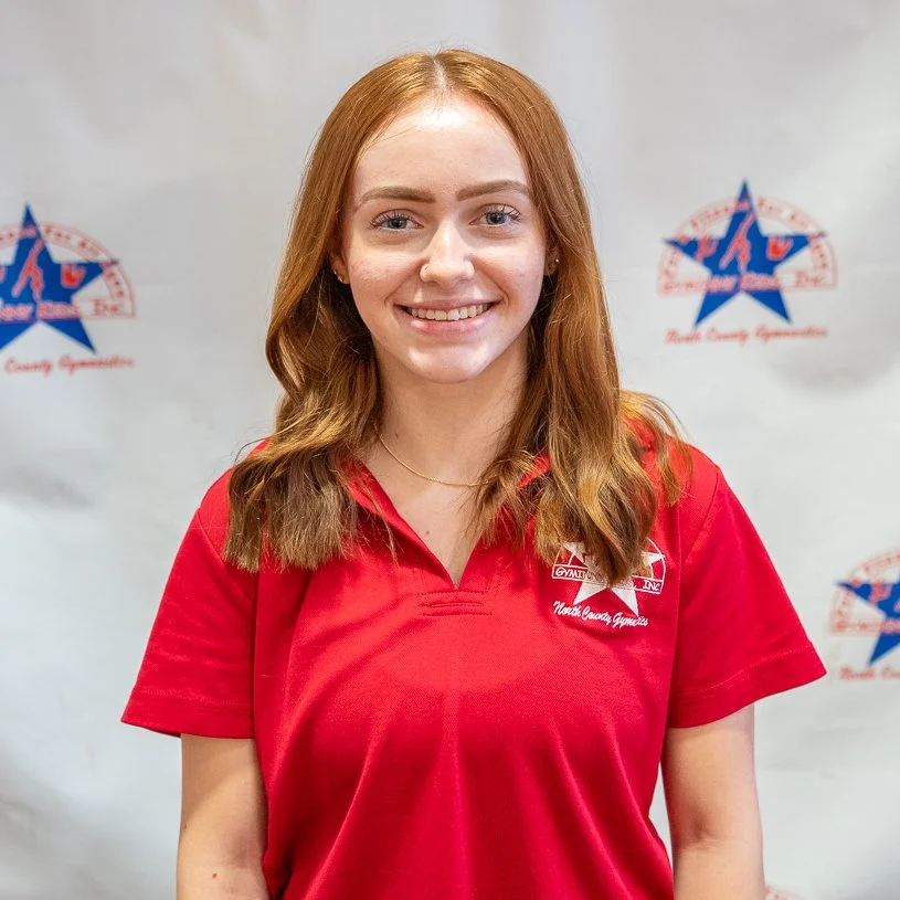 Young woman with red hair smiling, wearing a red shirt with a logo on it, standing in front of a backdrop with a star and text that reads "North County Gymnastics."