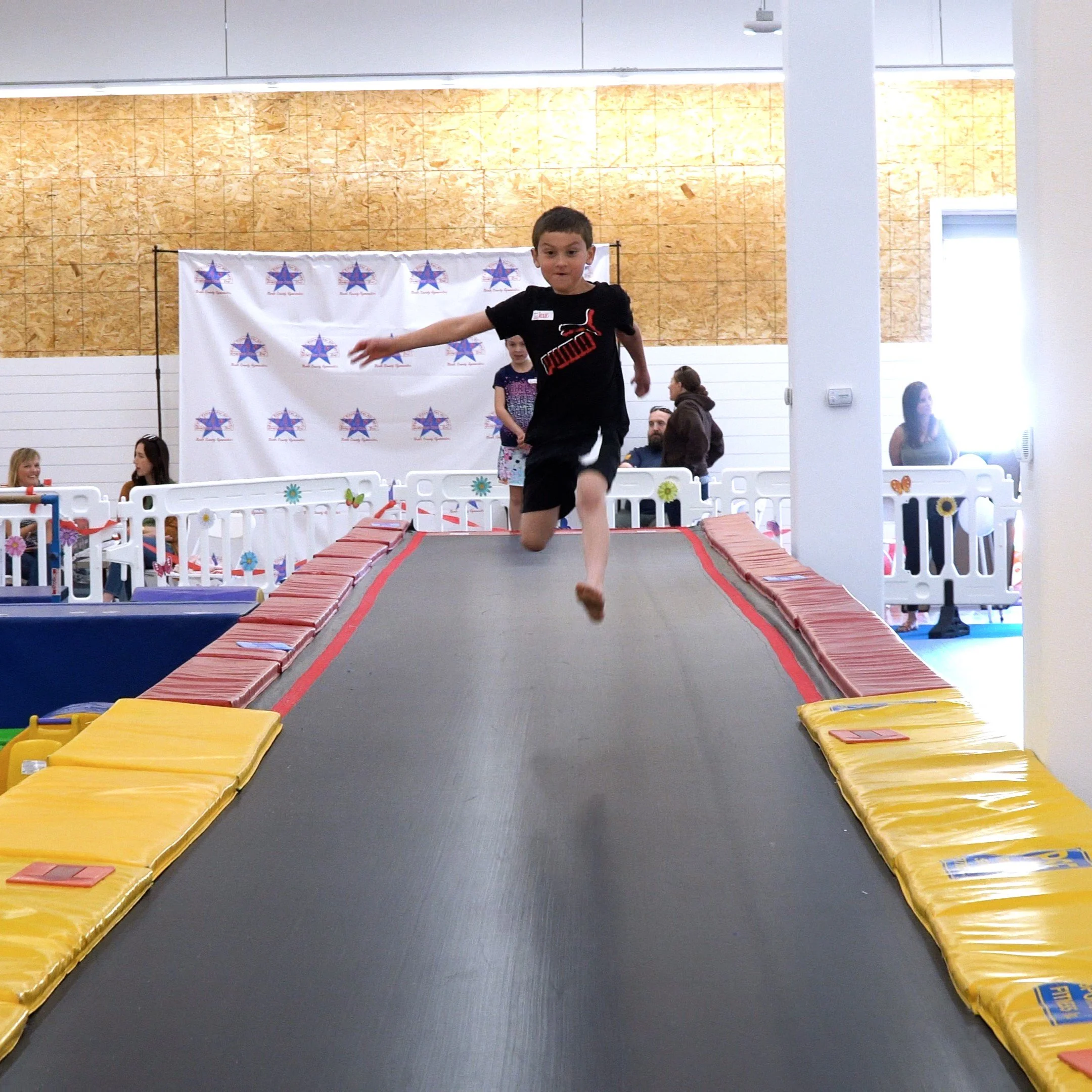 A young boy is jumping on a trampoline at an indoor recreational facility, with adults and children seated and standing in the background.