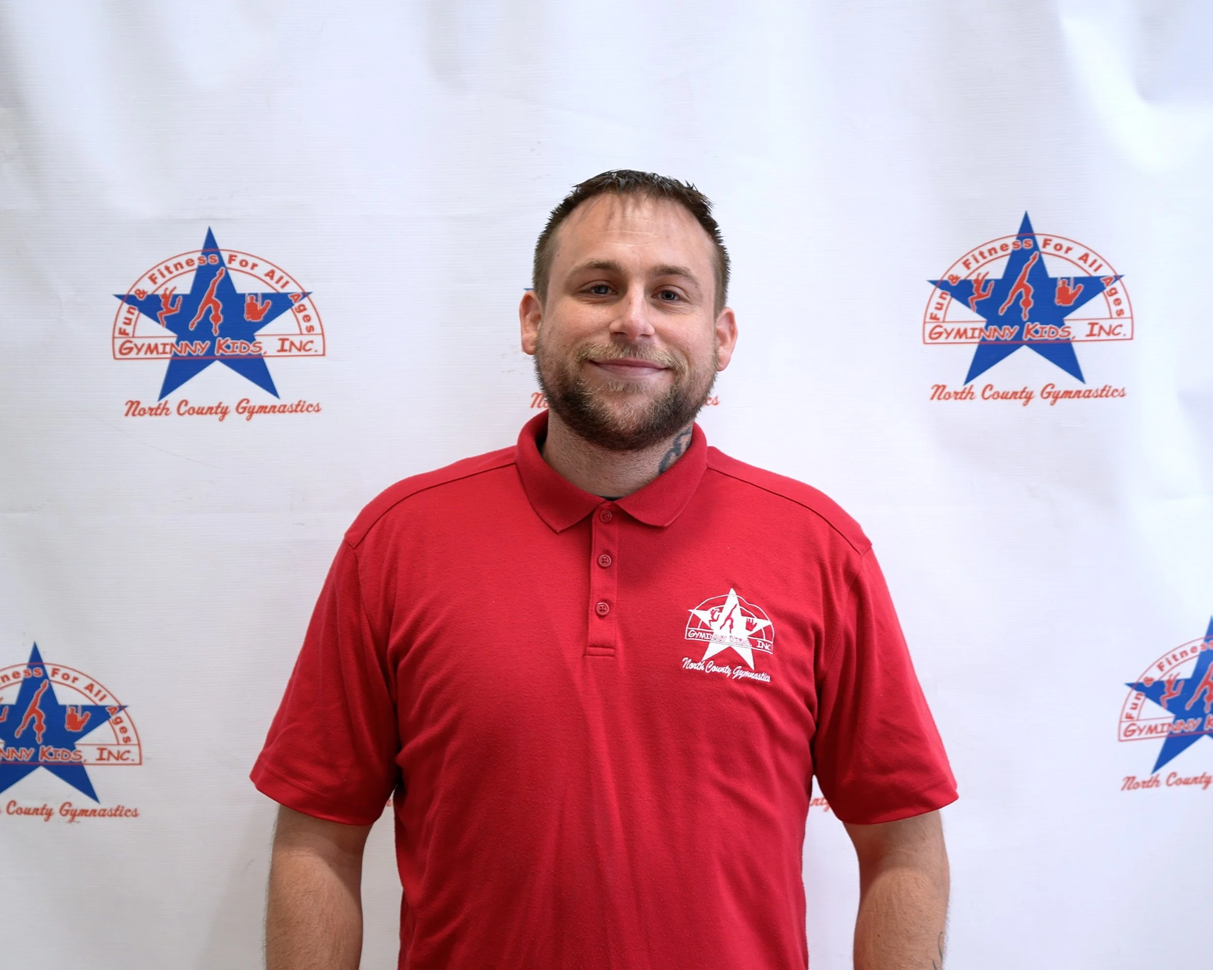 A smiling man with short hair and a beard standing in front of a backdrop with Gyminy Kids Inc. and North County Gymnastics logos, wearing a red polo shirt with the gym's logo.