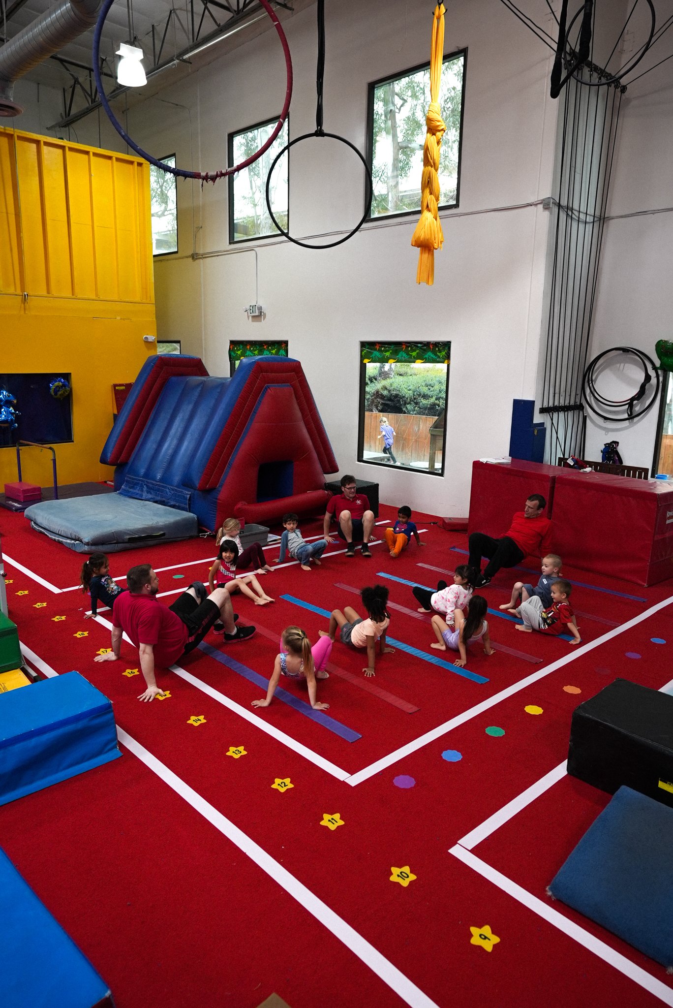 Children and instructors participating in a gymnastics class inside a gym. There is an inflatable slide, hanging gymnastics rings, and various colored mats and foam blocks on a red floor. Large windows and gym equipment are visible in the background.