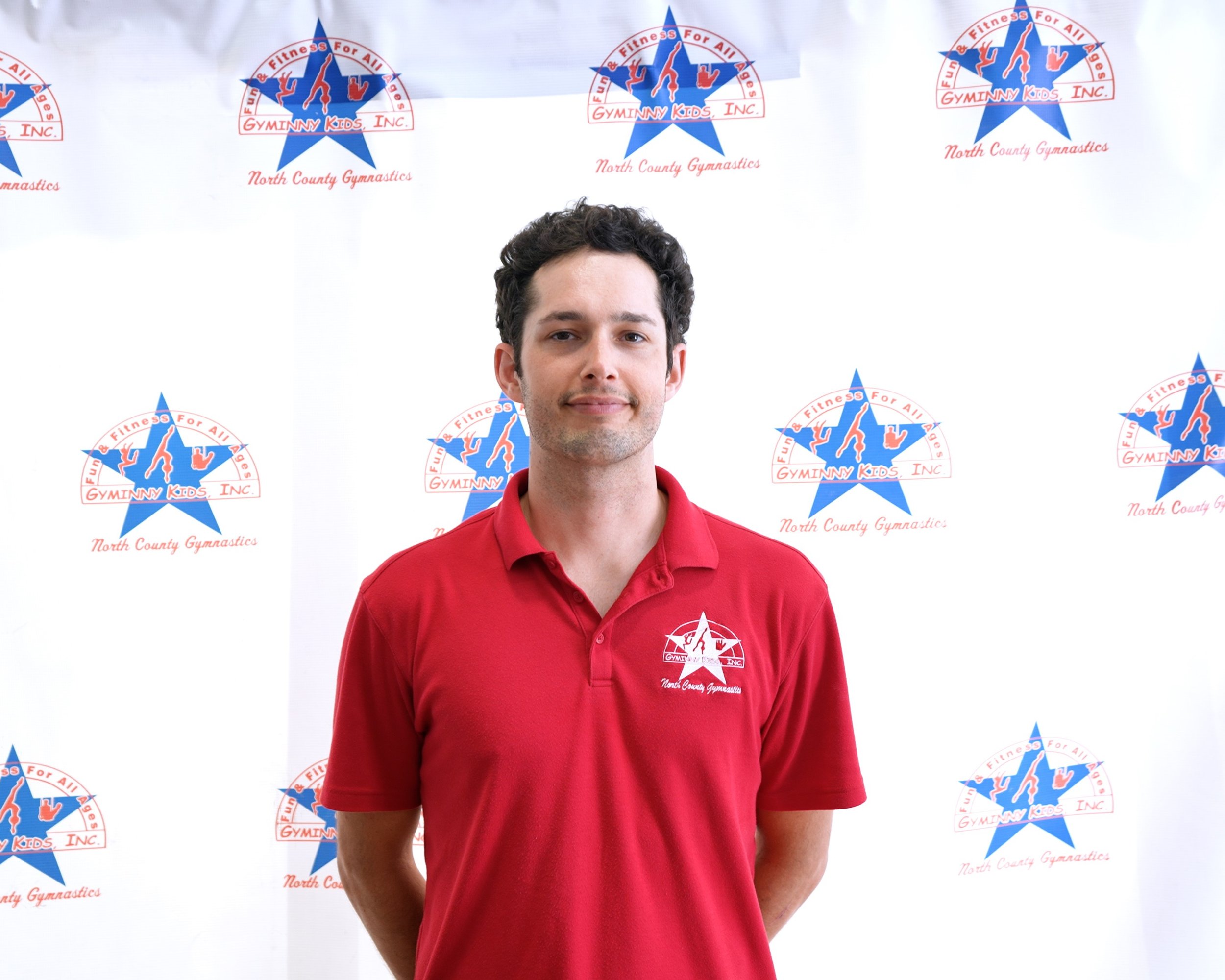 A man in a red polo shirt standing in front of a backdrop with gymnastic logos for North County Gymnastics and Gymnanny Kids, Inc.