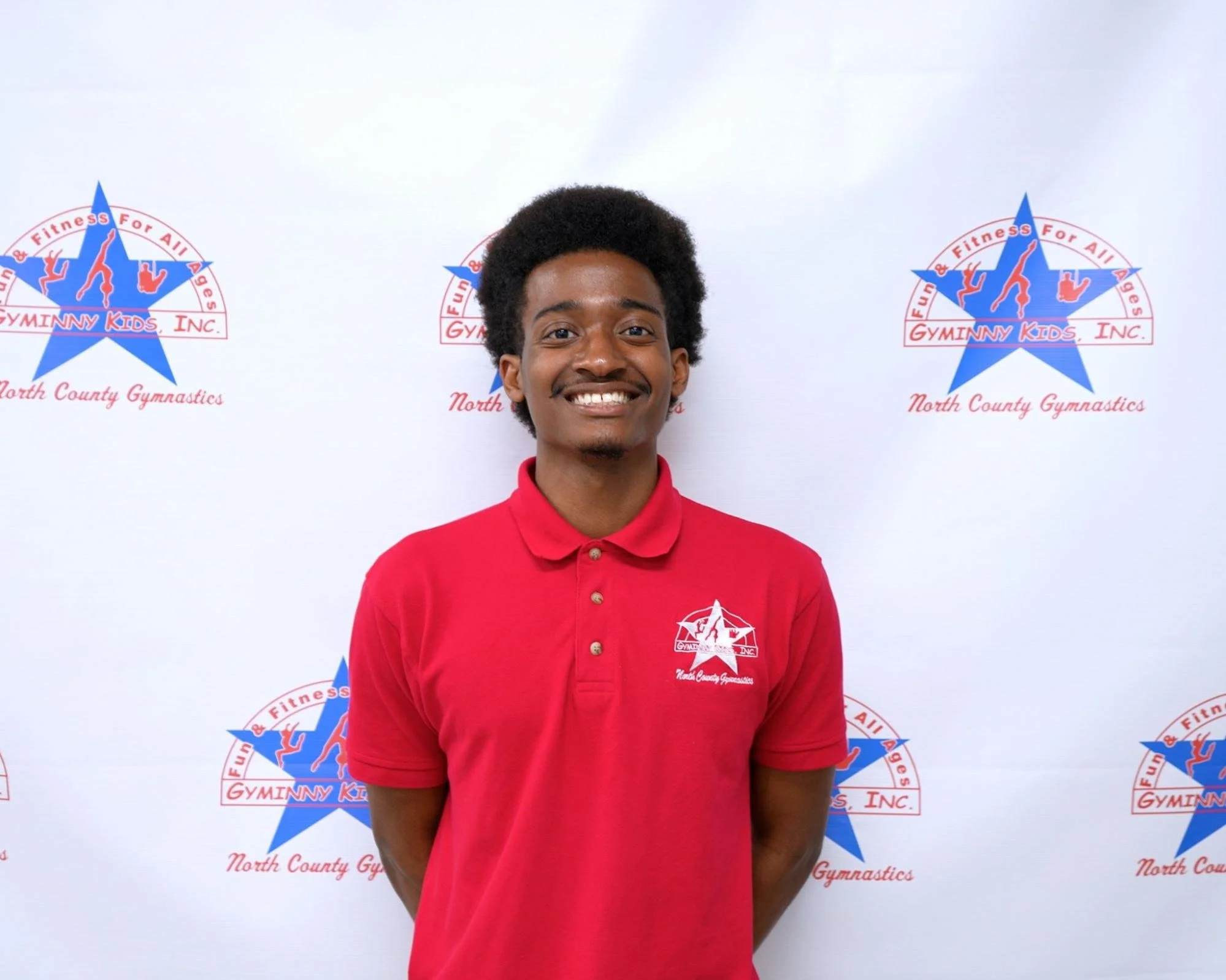 Young man wearing a red polo shirt with Gymniny Kids, Inc. logo, smiling and standing in front of a Gymniny Kids, Inc. North County Gymnastics backdrop.