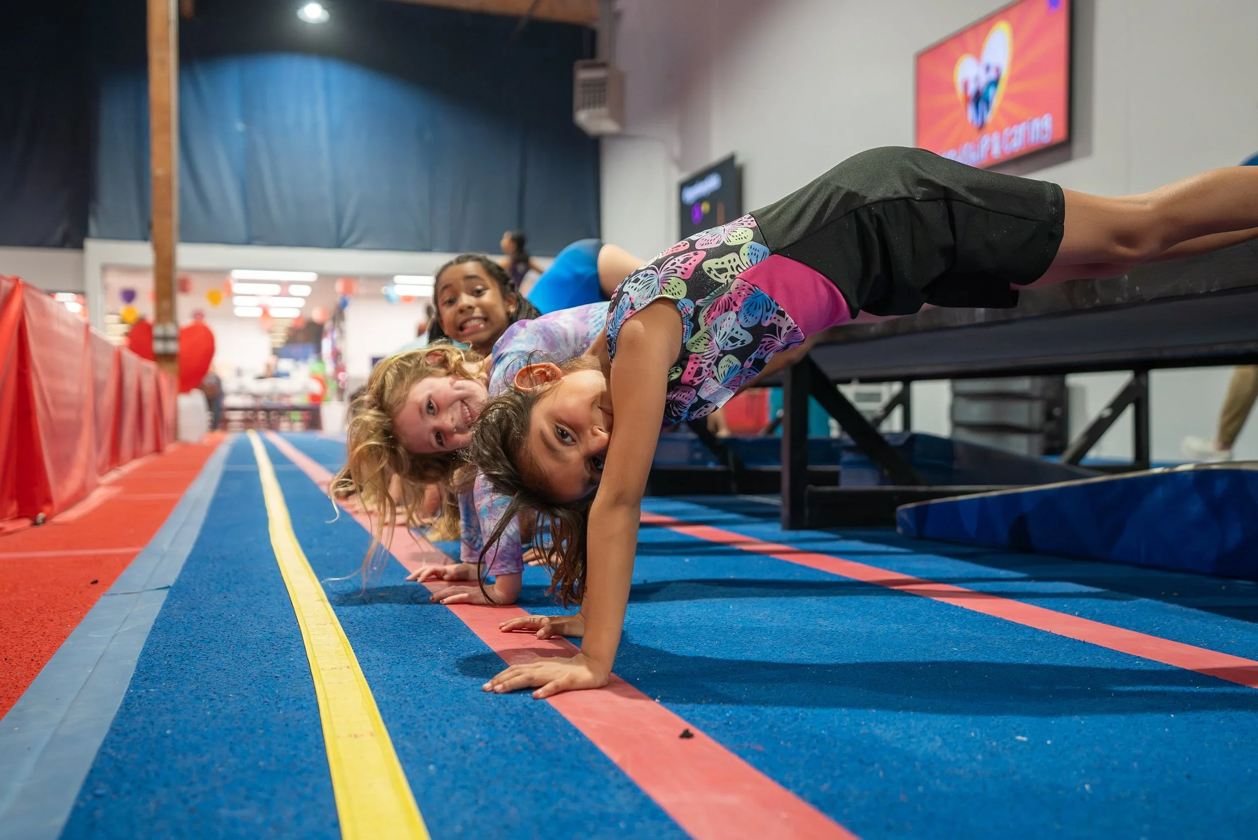 Children celebrating a birthday party with a decorated jungle theme. Kids sit around a table with a tiger-striped cake, balloons, and party favors, while a woman takes a photo of the children. The background features jungle animal murals and festive decorations.