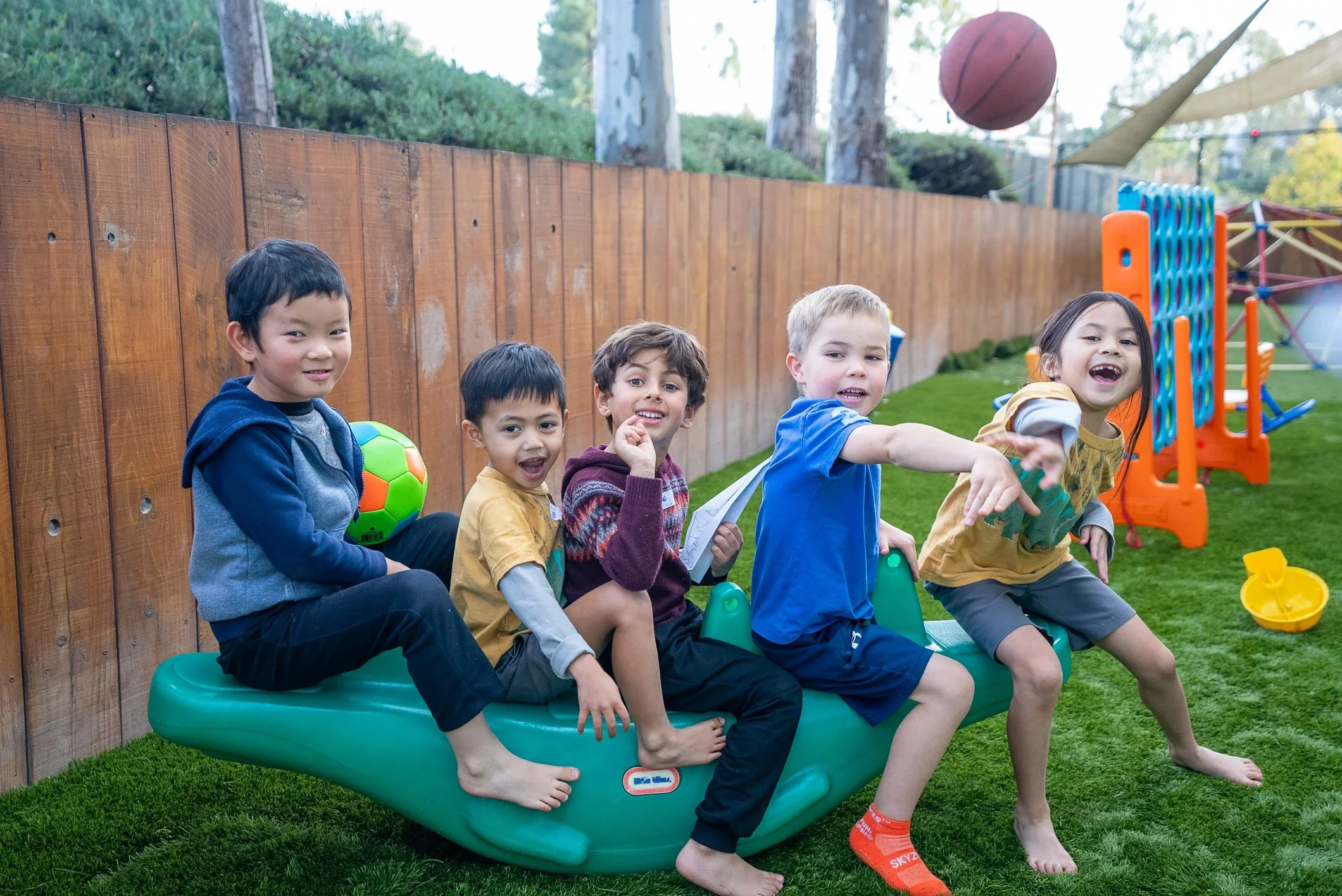 Five children sitting on a green plastic seesaw in a backyard with a wooden fence, playing with a ball and smiling.