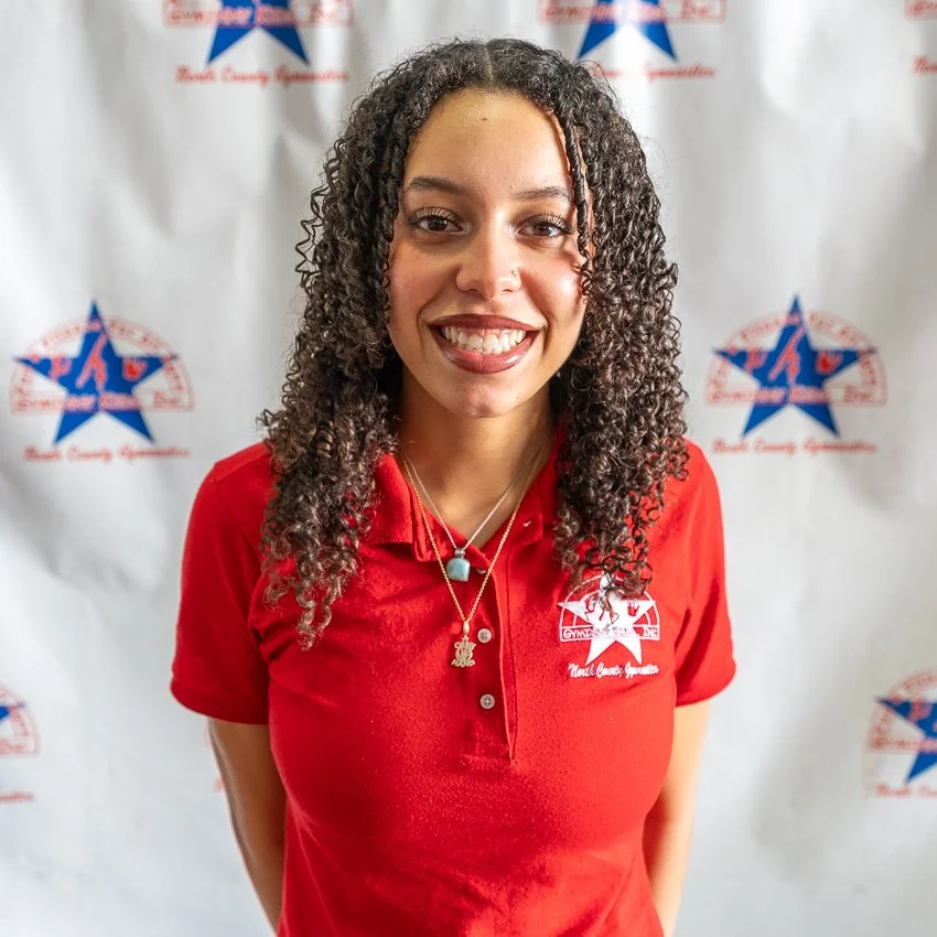 Young woman with curly hair smiling, wearing a red shirt with a logo, standing in front of a white backdrop with blue and red patriotic stars and text.