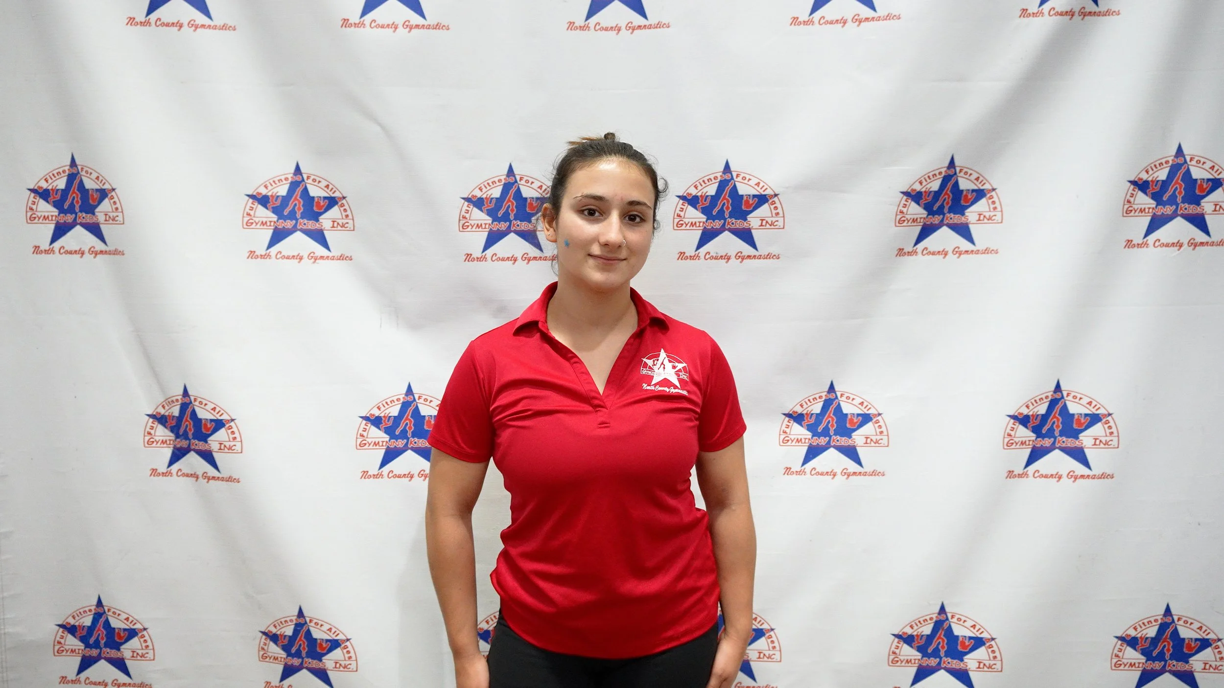 A young woman standing in front of a backdrop with the Gymnastics for All and North County Gymnastics logos, wearing a red polo shirt and black pants.