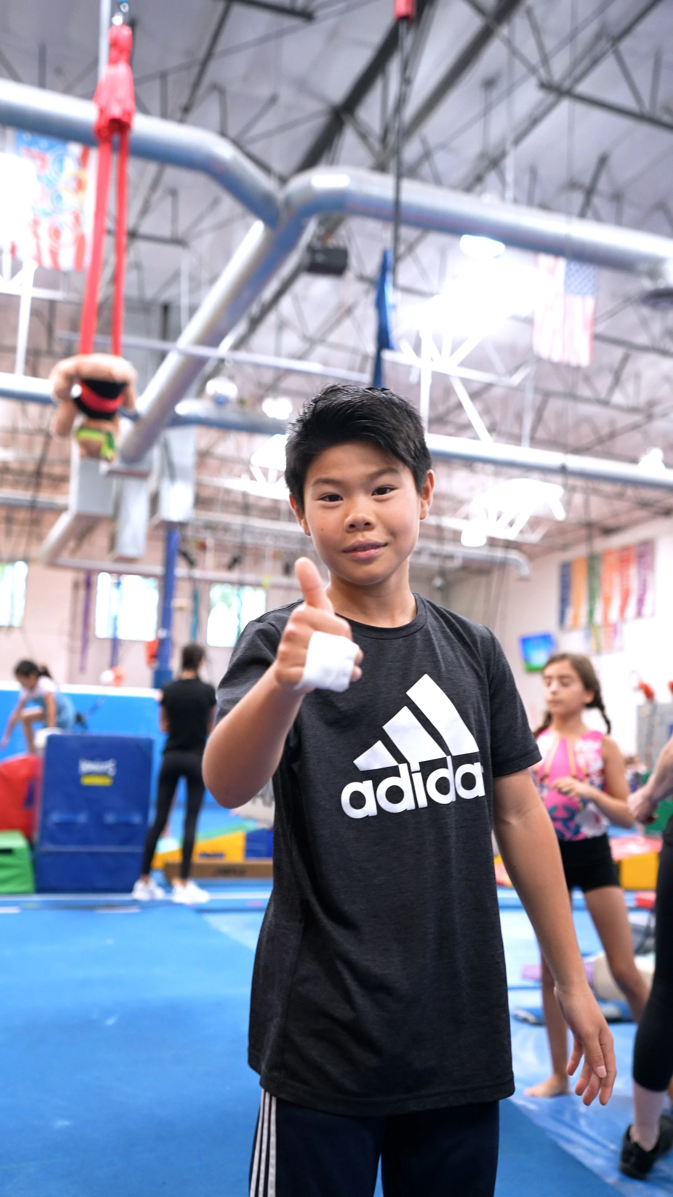 A young boy giving a thumbs-up in a gymnastics gym, with gymnastic equipment, hanging rings, and other children visible in the background.