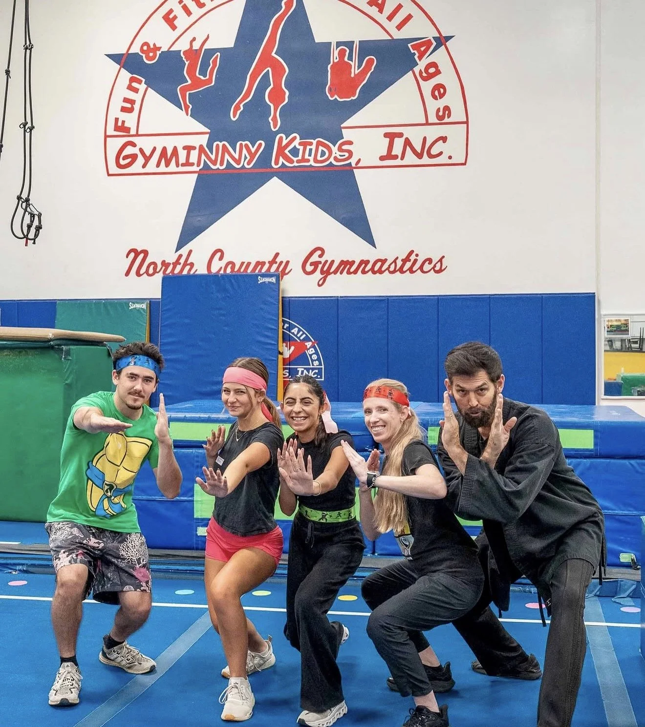 Gyminny Kids coaches strike playful ninja poses beneath the North County Gymnastics wall logo.
