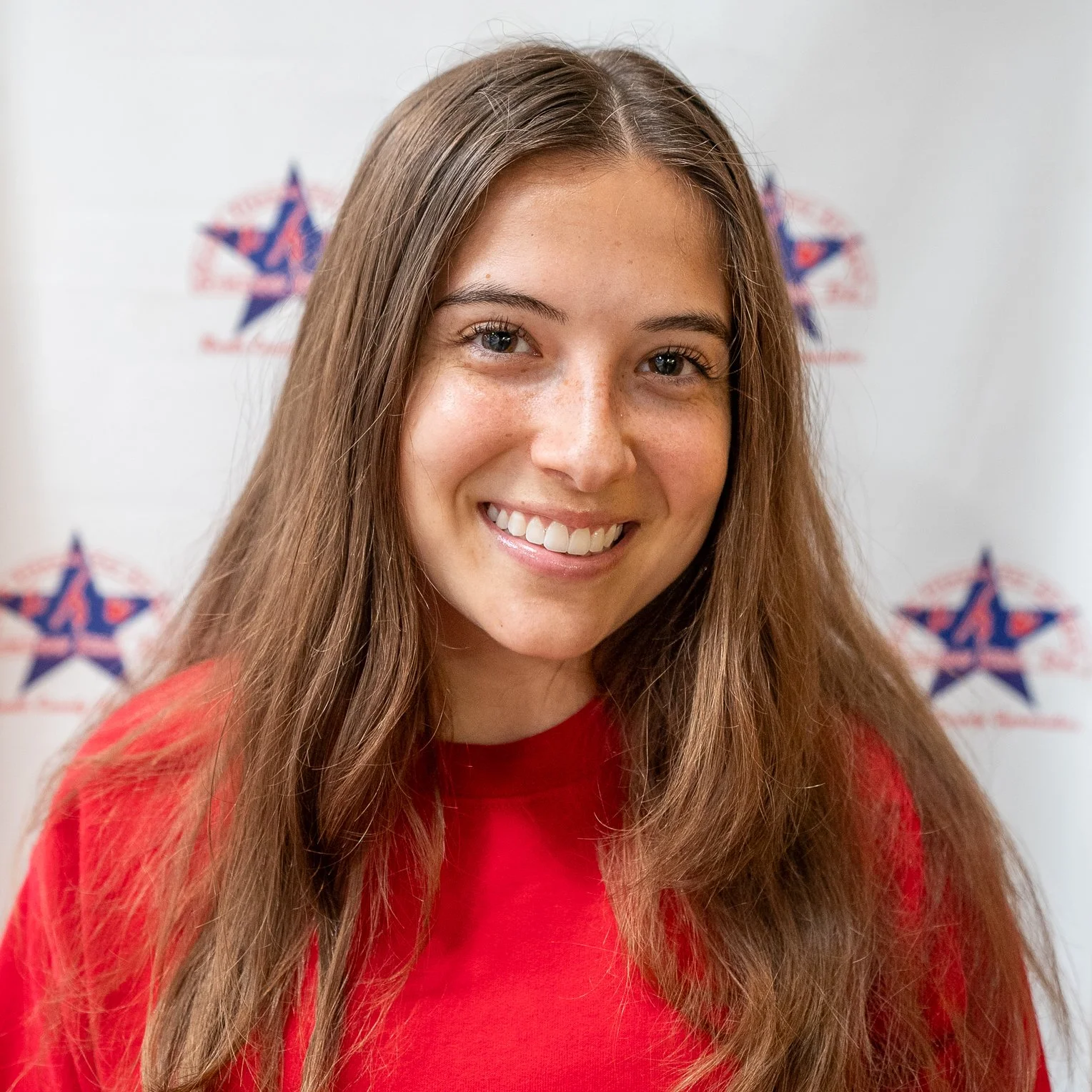 Portrait of a smiling young woman with long brown hair wearing a red shirt standing in front of a white background with a star logo.