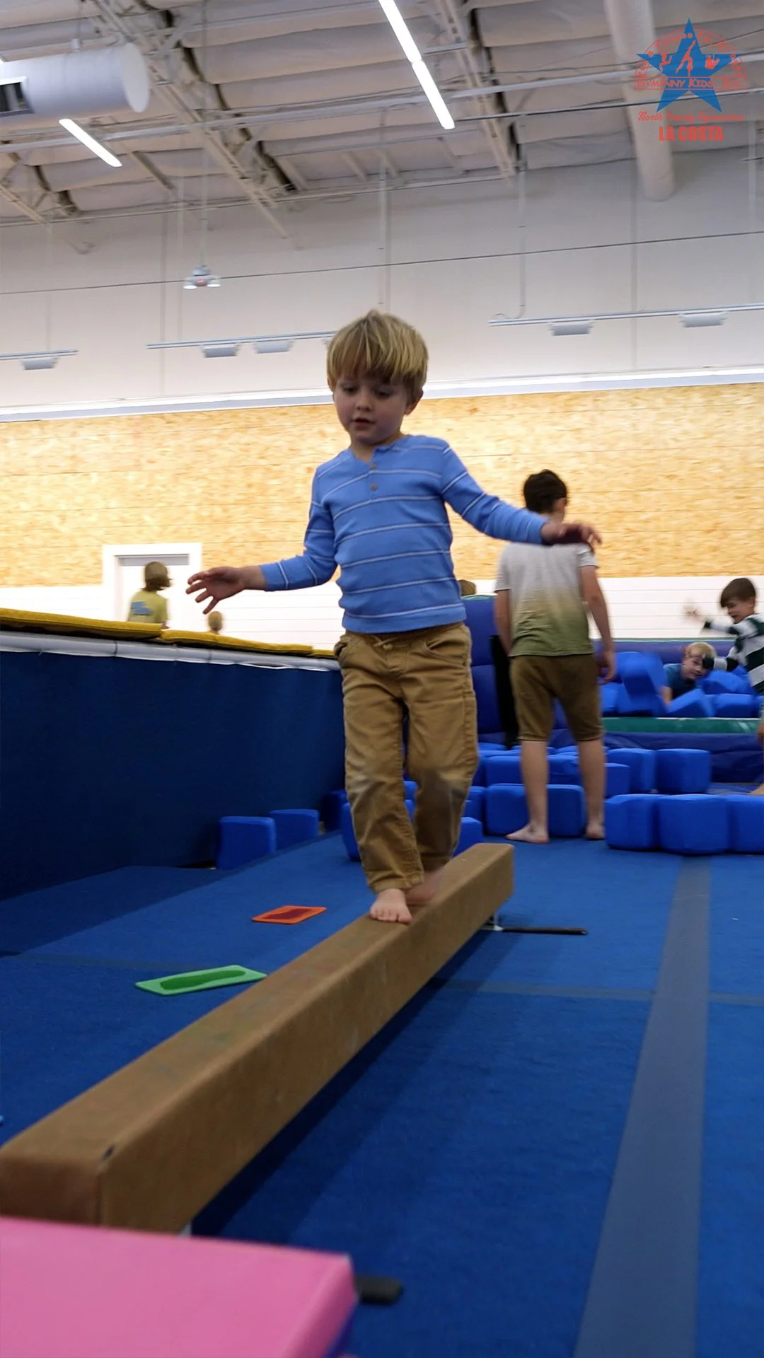 A young boy with blond hair balancing on a wooden beam at an indoor gymnastics facility, with other children in the background playing with blue foam blocks.