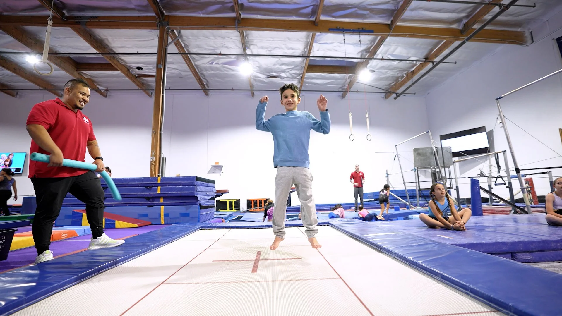 Children and adults in a gymnastics gym, with a boy jumping on a trampoline, surrounded by gymnastic equipment and other kids practicing.
