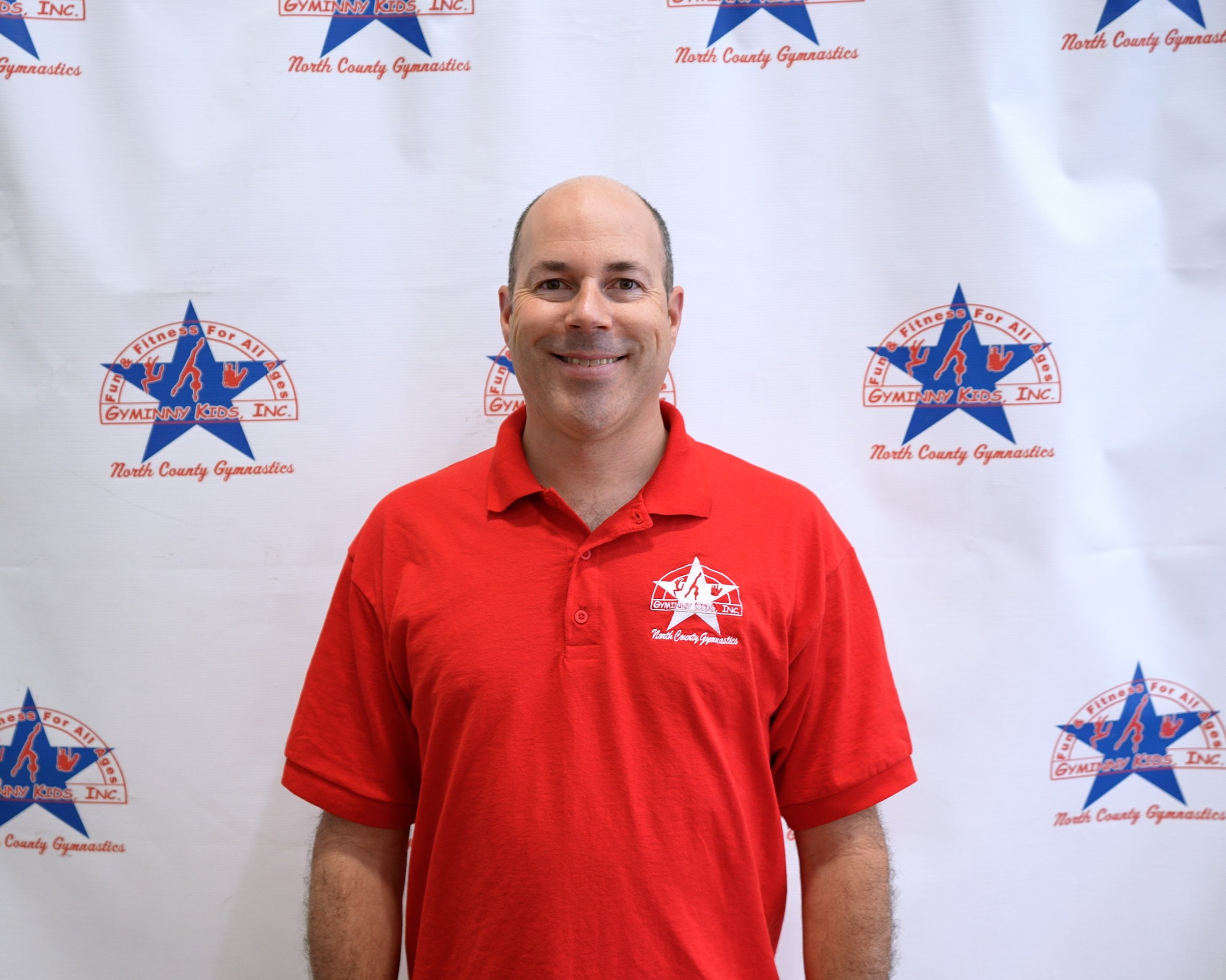 Man smiling in front of a backdrop with Gymnny Kids Inc. and North County Gymnastics logos, wearing a red polo shirt with the same logo.