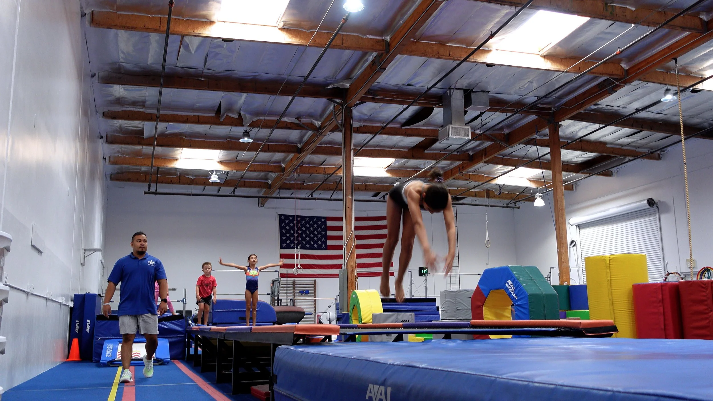 A gymnastics training facility with a girl mid-air doing a somersault on a trampoline, a coach walking nearby, and other children practicing activities in the background. The American flag hangs on the wall.