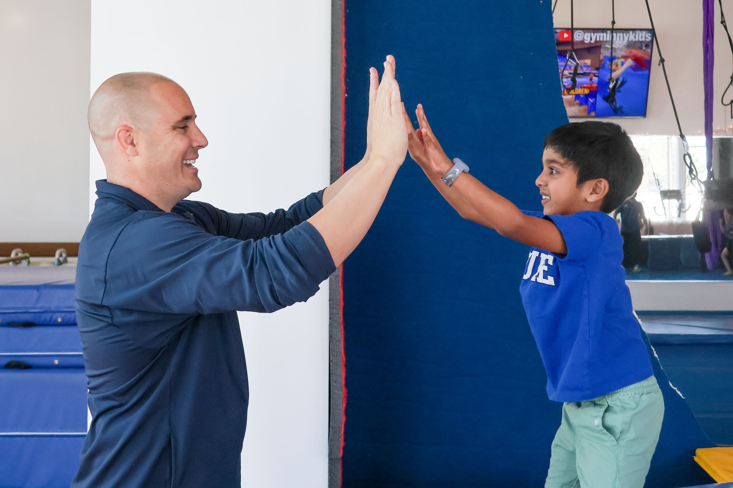 Four people standing in an indoor trampoline park, all wearing red shirts and black or khaki pants, smiling at the camera. The background shows colorful trampolines and a sign that reads 'Spring Break & Summer Camp'.