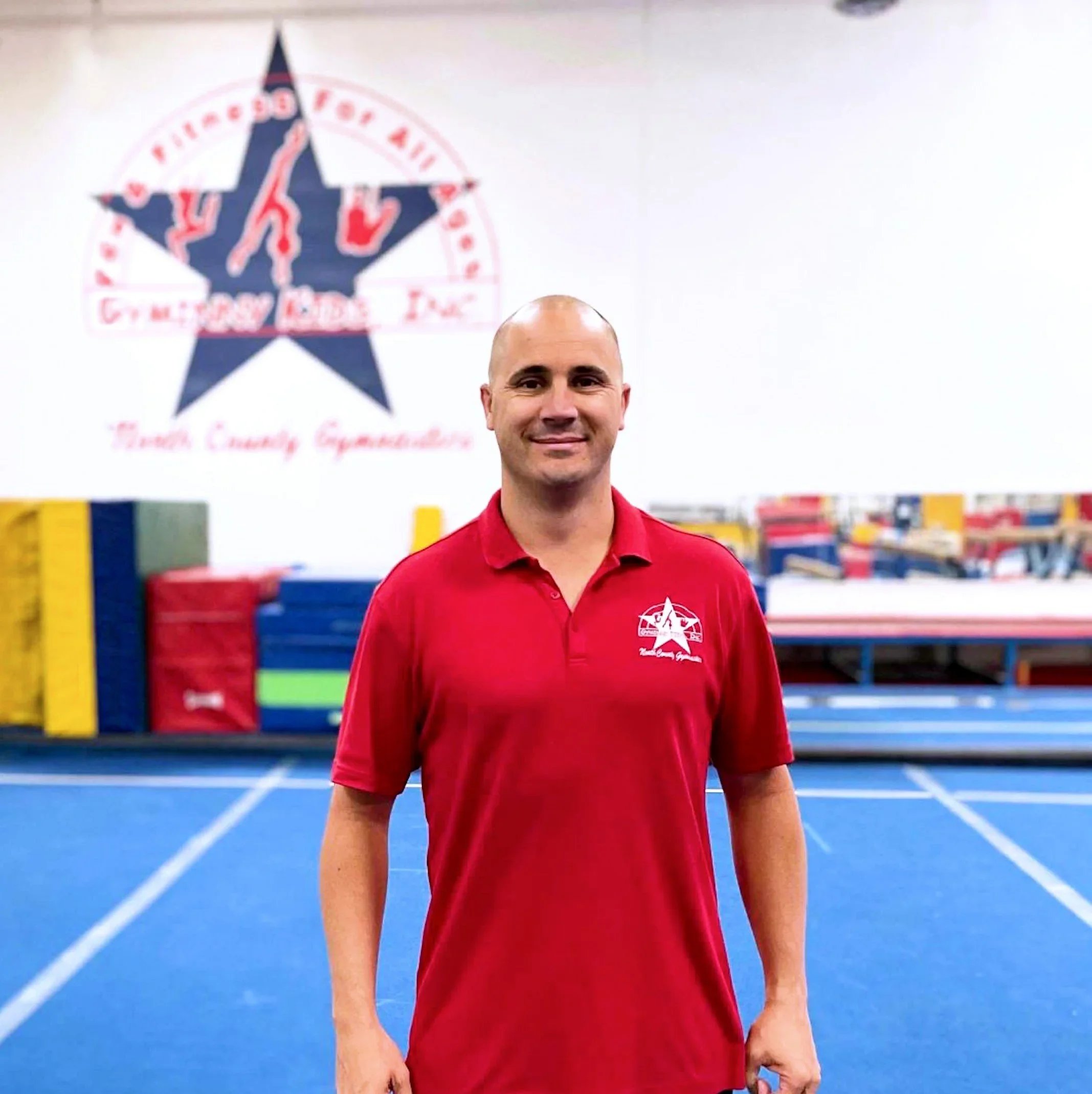 Four people standing in an indoor trampoline park, all wearing red shirts and black or khaki pants, smiling at the camera. The background shows colorful trampolines and a sign that reads 'Spring Break & Summer Camp'.