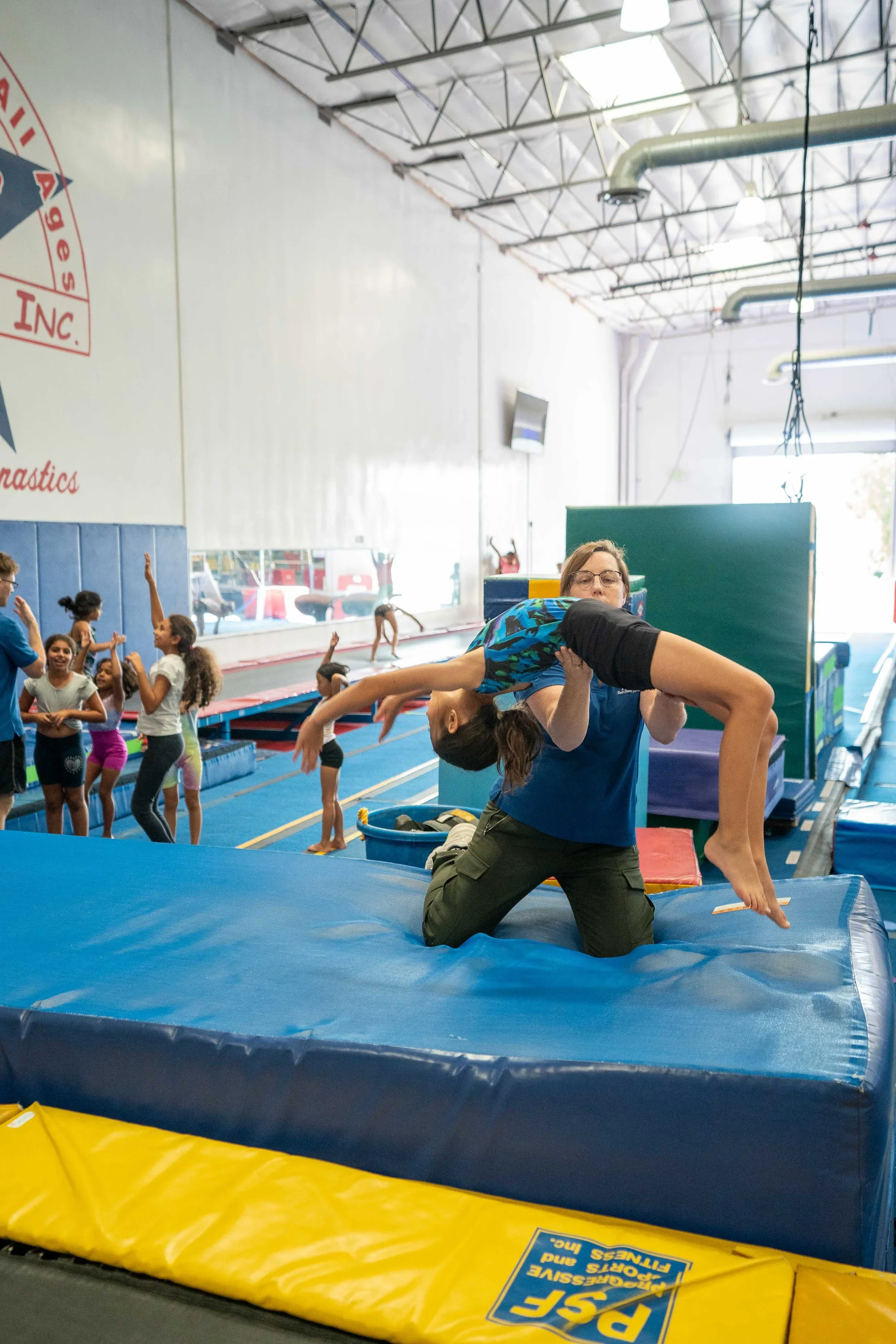 A young gymnast is being supported by an instructor during a backbend on a padded gymnastics mat inside a gym facility. In the background, a group of children are waiting, some with their hands raised, in a line along the wall.