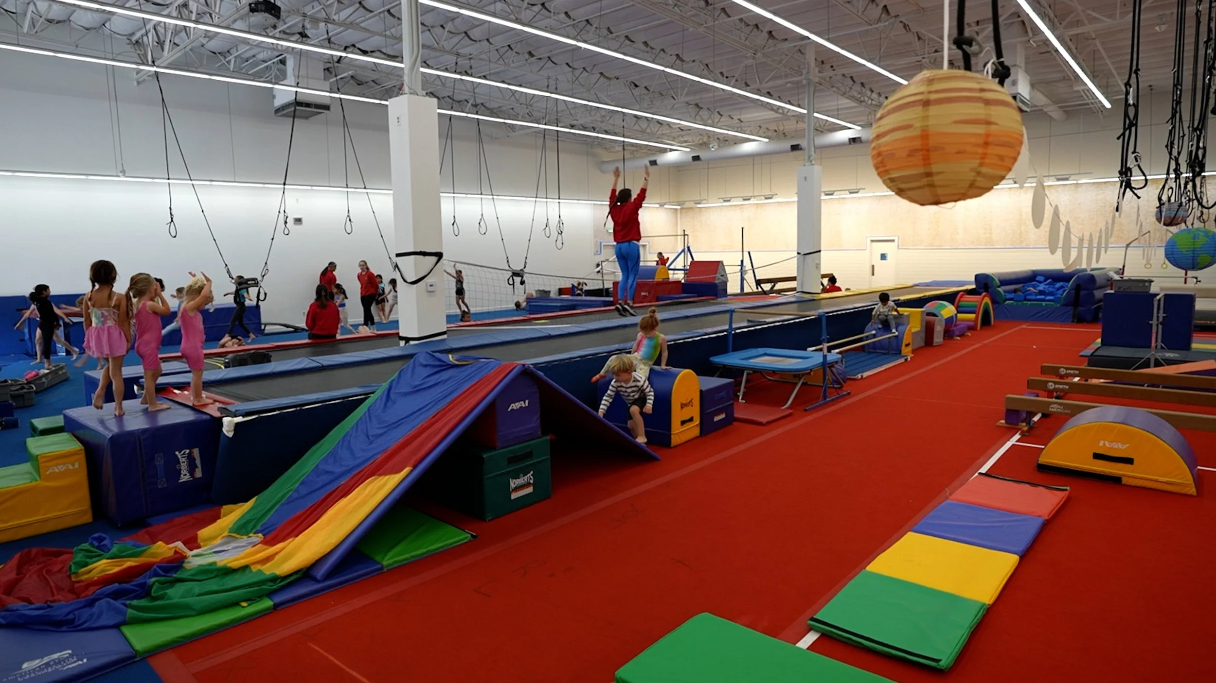 Children and adults in an indoor trampoline park with various trampolines, padded obstacle courses, and colorful play equipment.