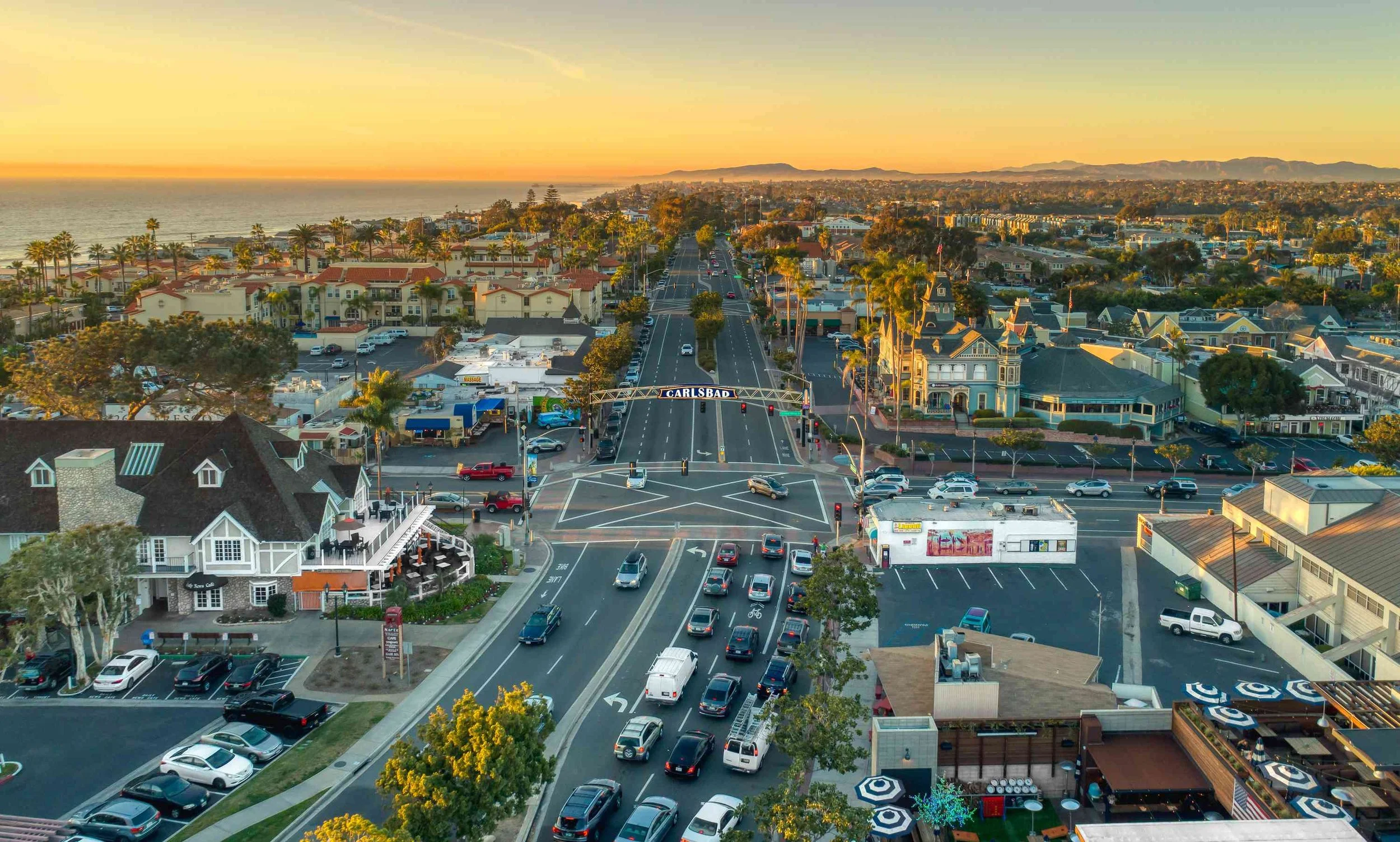 Aerial view of Carlsbad, California, during sunset showing a street with cars, buildings, and palm trees with the ocean and mountains in the background.