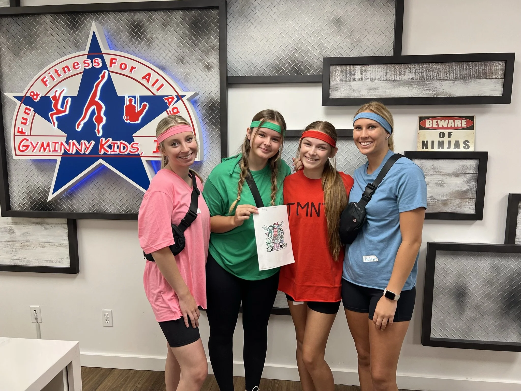 Gyminny Kids staff members in colorful headbands pose beneath the gym's 'Fun and Fitness for All Ages' sign.