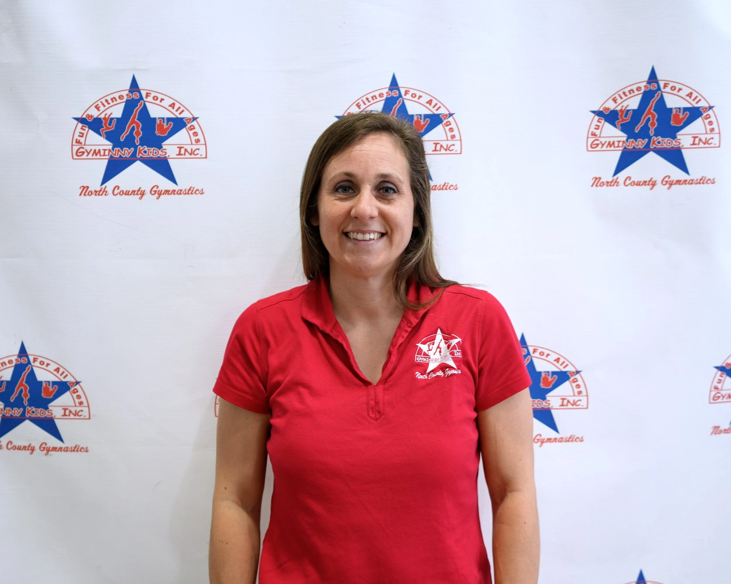 Women smiling in front of a Gymnanny Kids Inc. North County Gymnastics backdrop, wearing a red shirt with the gym's logo.