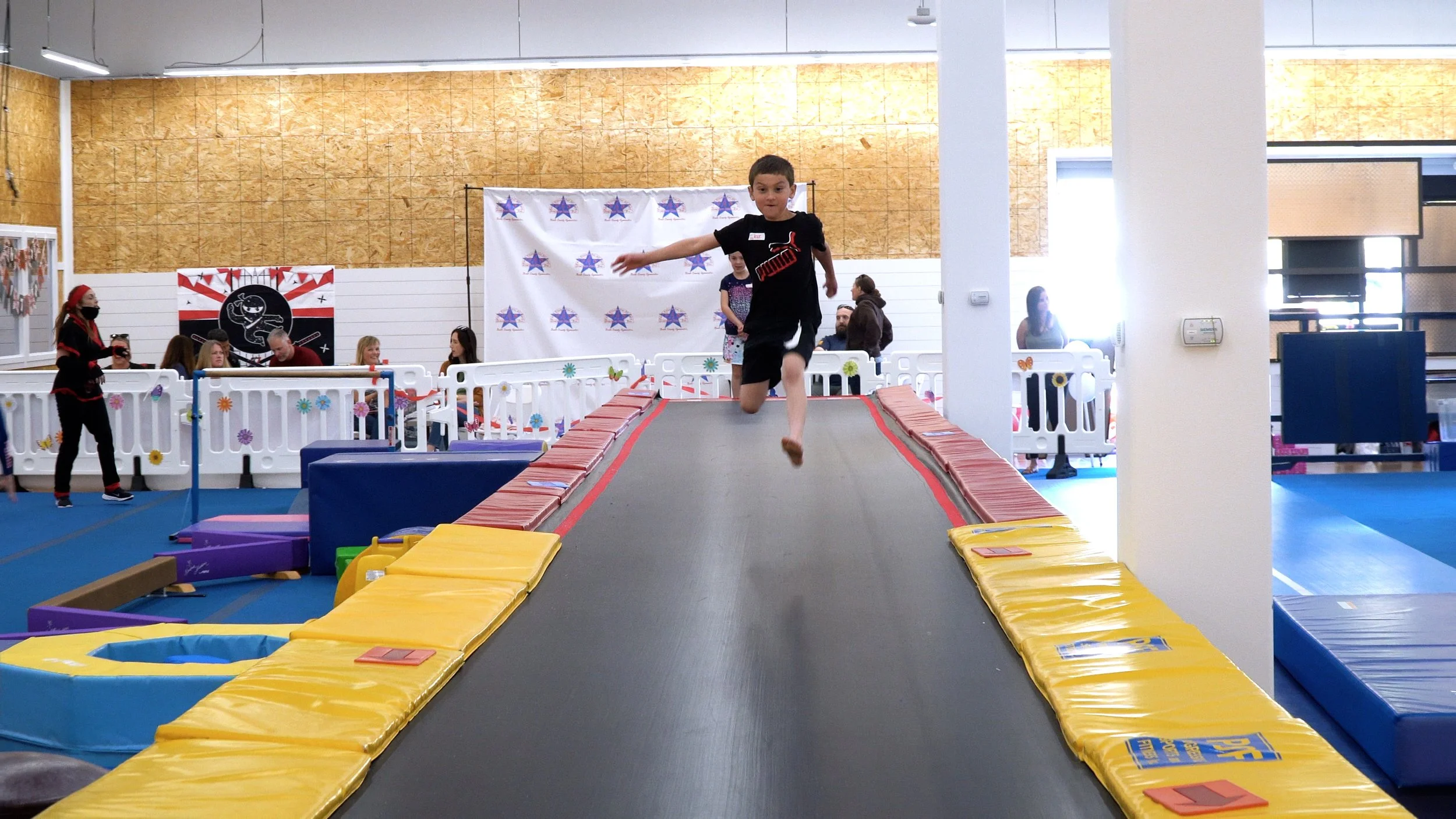 Child jumping on a trampoline at an indoor play center with colorful padded edges and foam blocks nearby, adults and children in the background, festive decorations, and a wall with patriotic and superhero-themed posters.