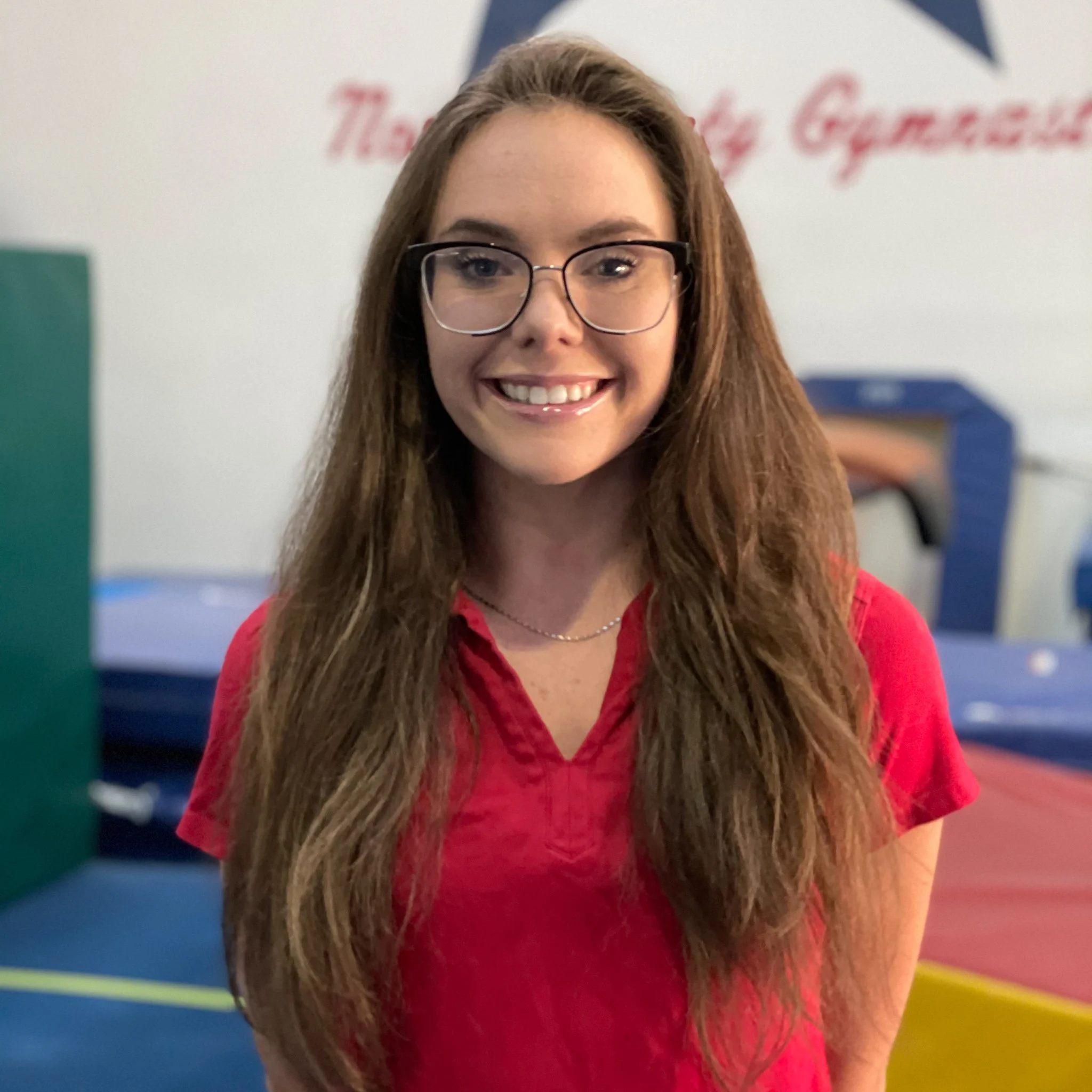 A young woman with long brown hair, wearing glasses and a red shirt, smiling in an indoor gymnastics facility.