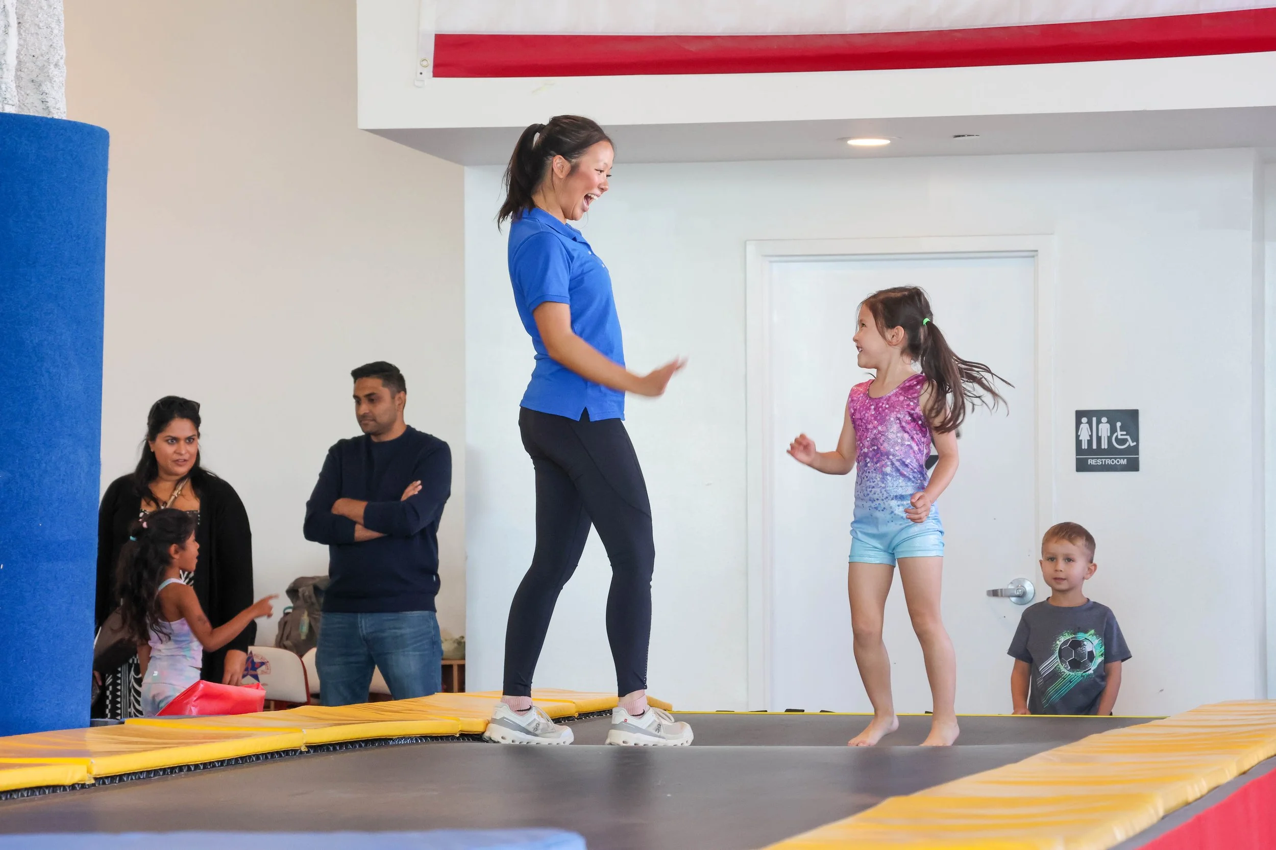 A young boy hangs upside down from a gymnastics bar in a gym, smiling at the camera, with a banner in the background reading 'Breaking Break & Summer'.