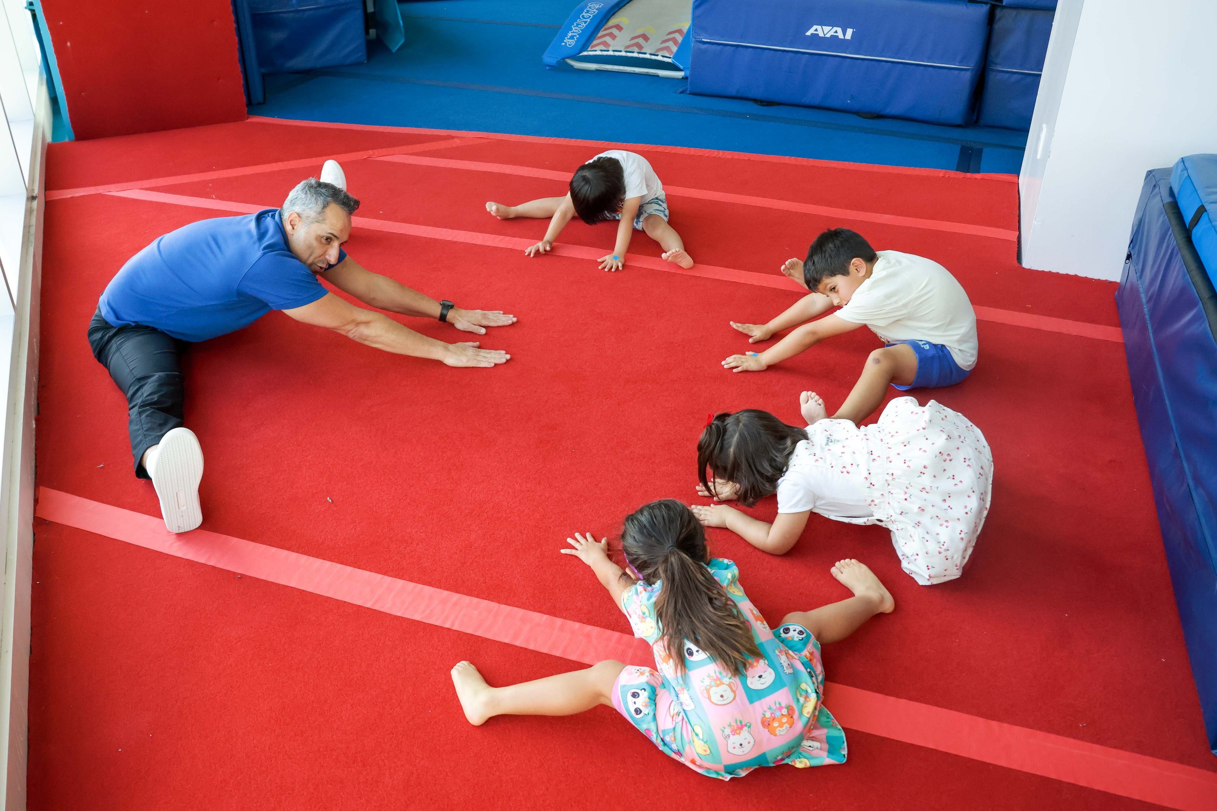 Young girl with curly hair smiling, sitting on a gymnastics vaulting horse at an indoor gym, with other children and gymnastic equipment in the background.