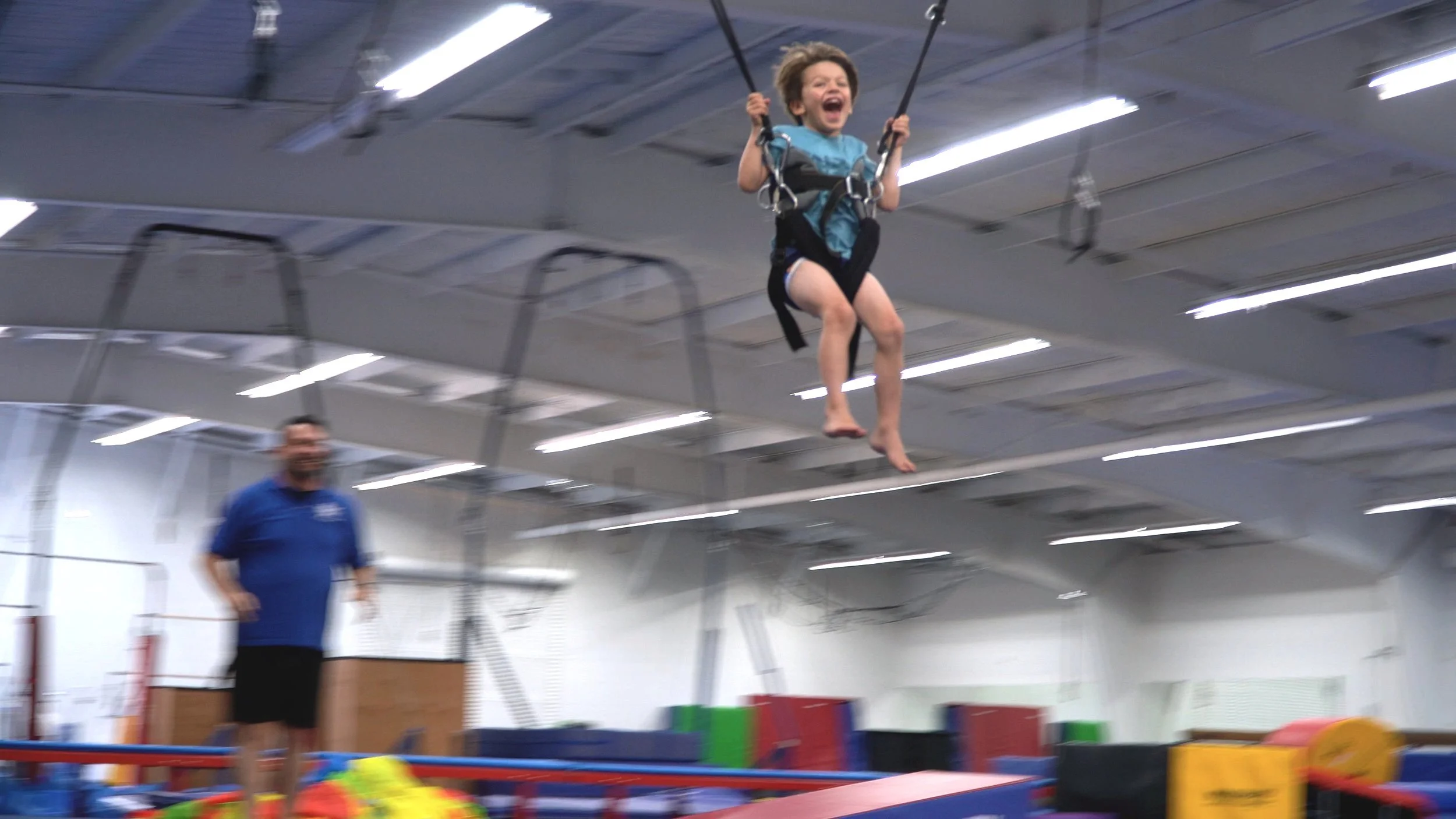 A young girl laughing as she swings on a trapeze in an indoor gym, with an adult instructor watching nearby.
