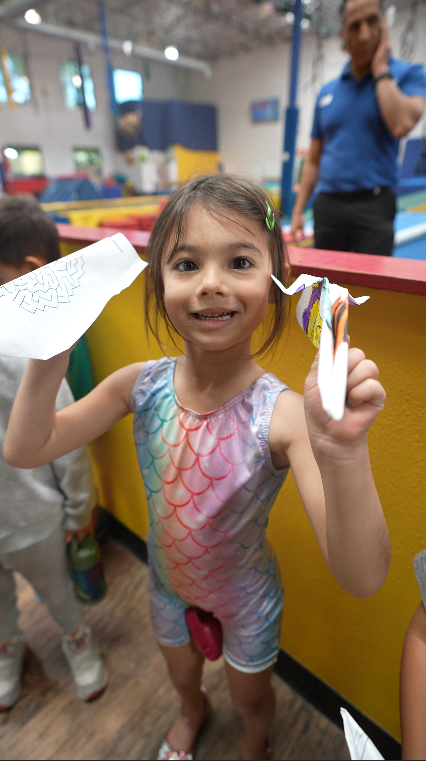 Young girl smiling and holding paper craft in a colorful indoor play area, with a man in a blue shirt in the background.