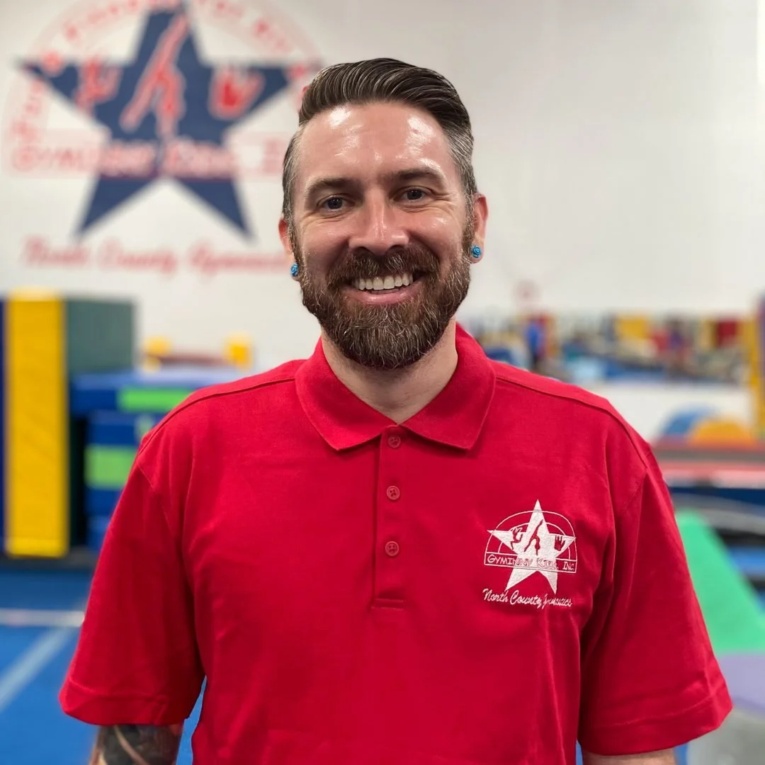 A smiling man with a beard, wearing a red shirt with a logo that reads 'Gymnastic Kids Inc. North County, Indiana,' standing in a gymnastics training facility with colorful equipment in the background.