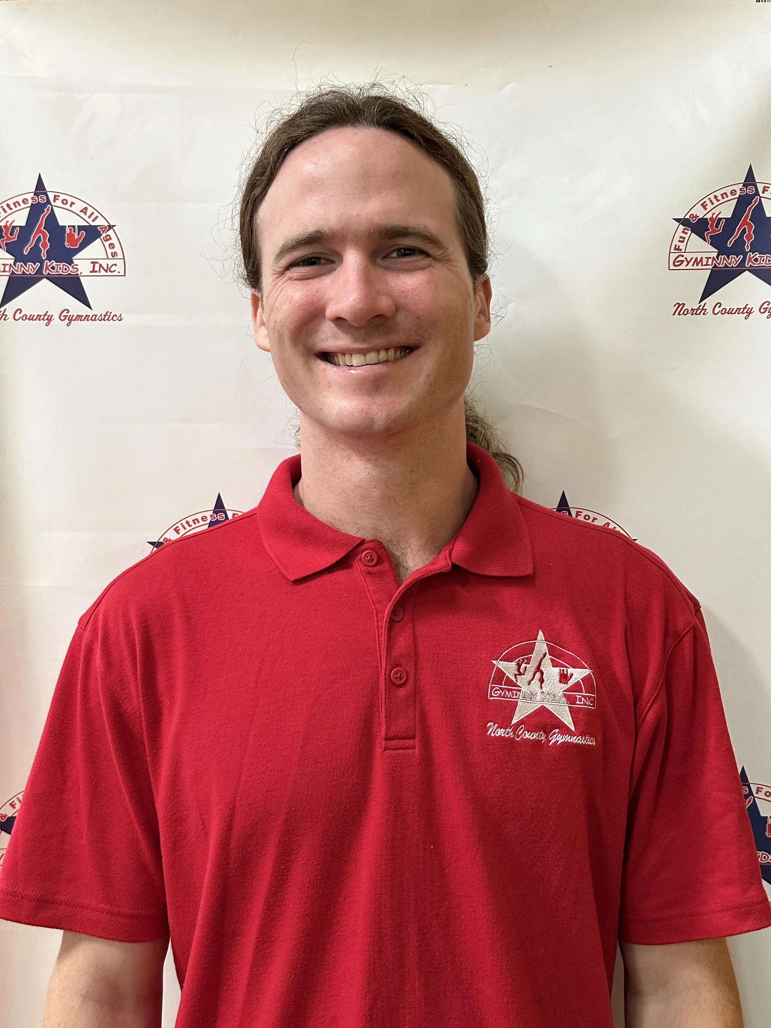 A smiling man with long hair tied back, wearing a red collared shirt with the Gymnanny Kids logo, standing in front of a white backdrop with gymnastic-themed logos.