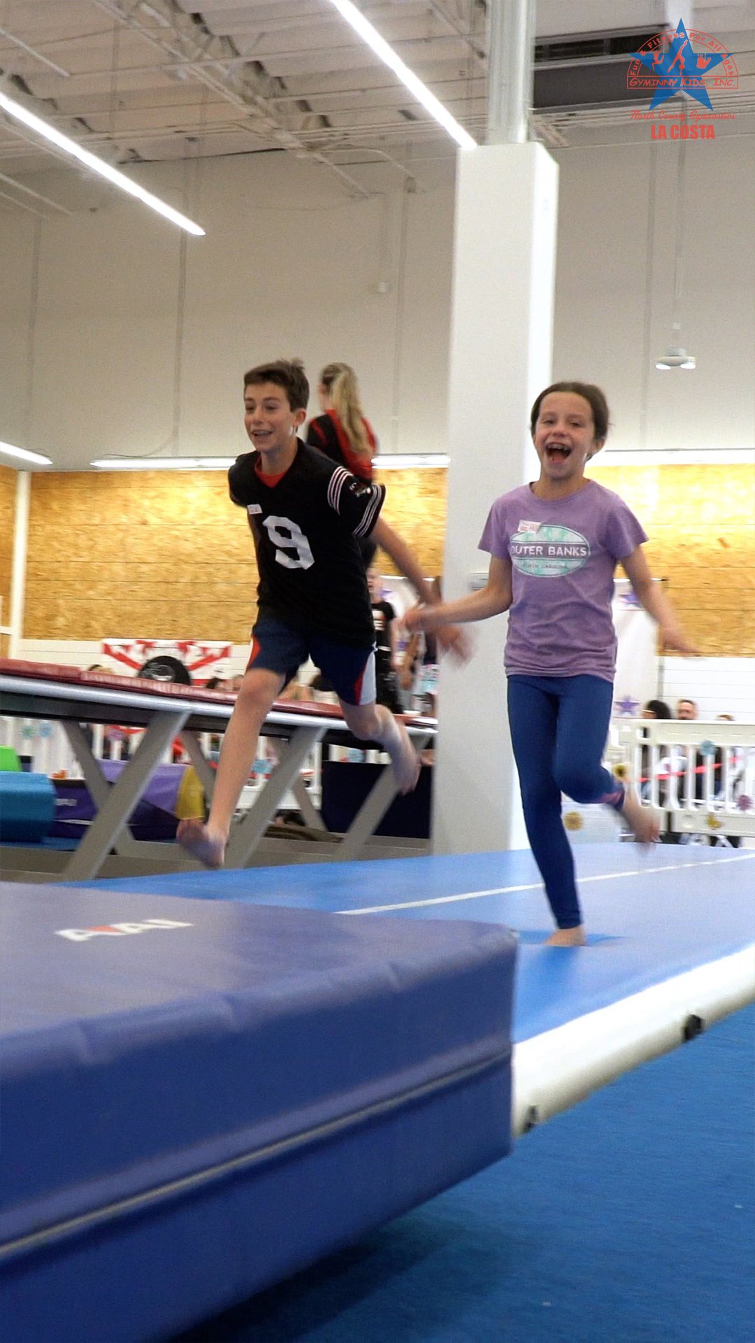 Two kids, a boy and a girl, are running and jumping on a gymnastic trampoline at an indoor gymnasium, smiling and enjoying.