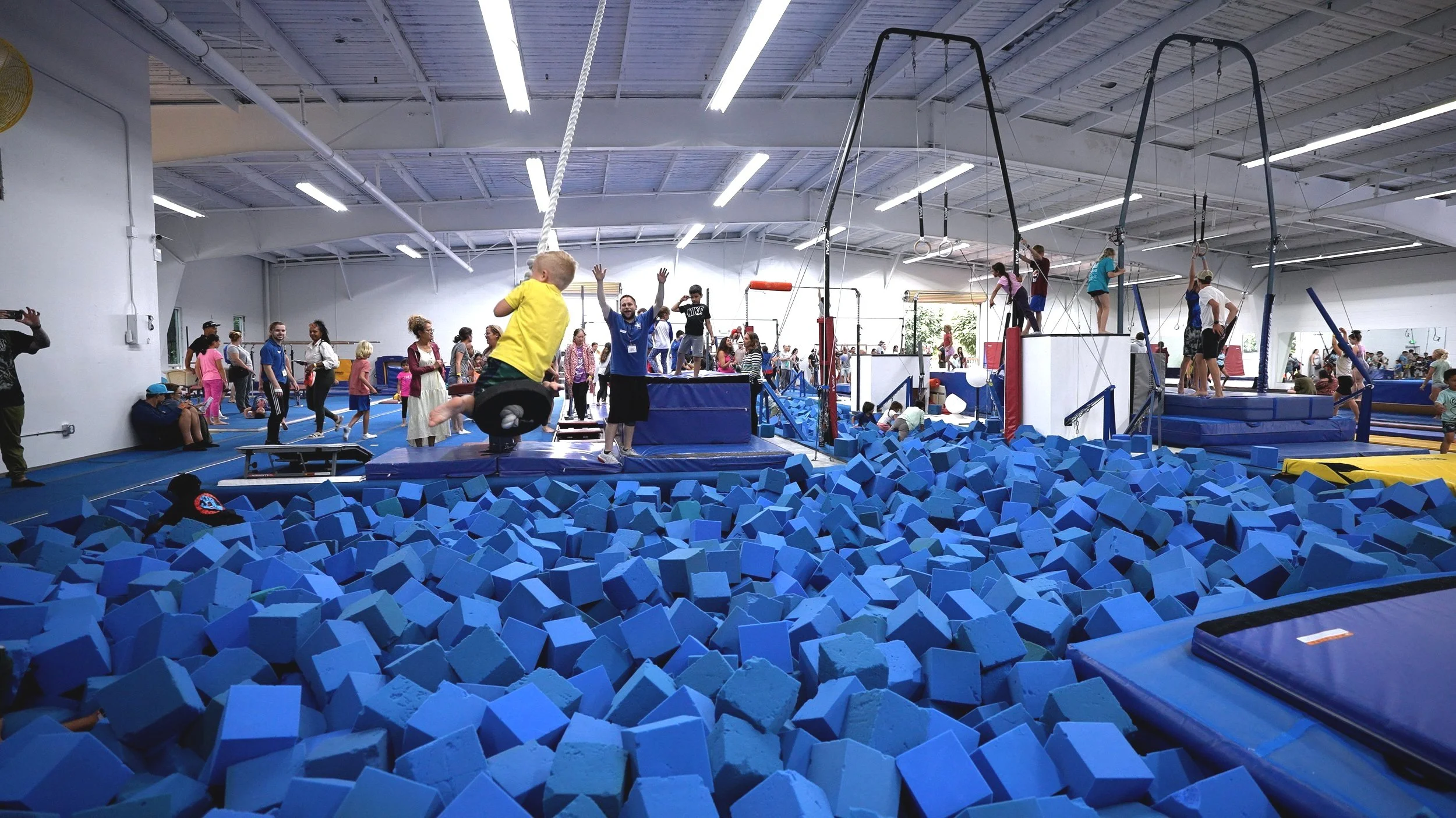 Indoor trampoline park with blue foam pit and children playing on various trampolines and climbing equipment.