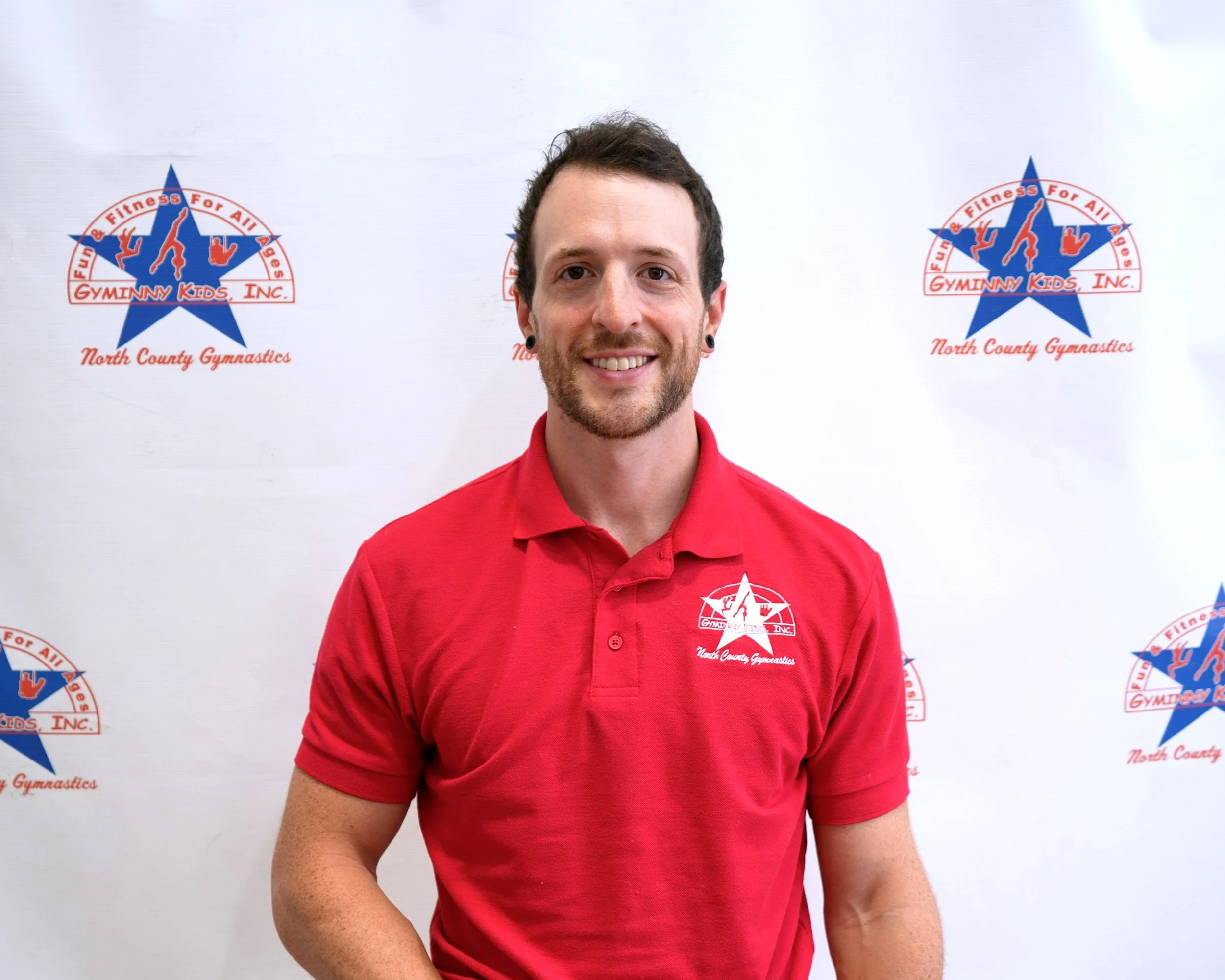 A man in a red polo shirt standing in front of a white backdrop with a gymnastics team logo. He has short dark hair, a beard, and earrings, and is smiling at the camera.