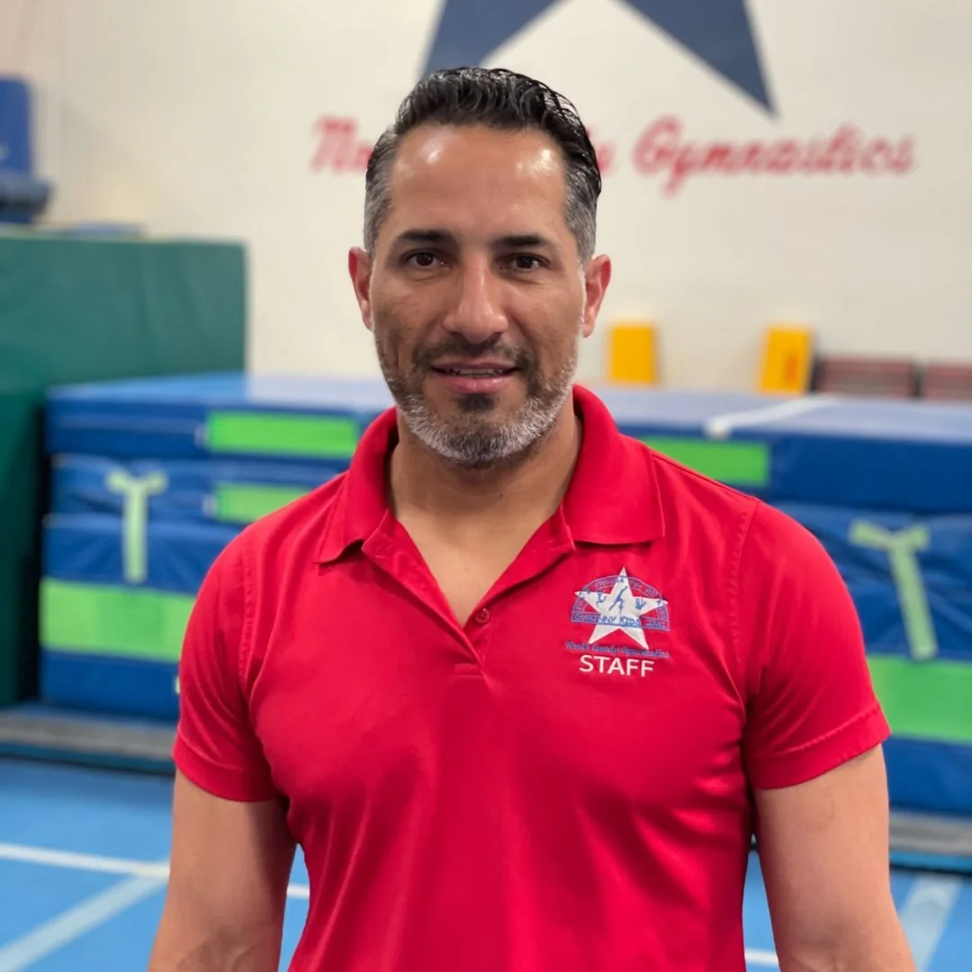 A man with dark, slicked-back hair and a beard wearing a red polo shirt with a staff logo, standing inside a gymnasium with blue and green sports mats and a wall with a large star and red text in the background.