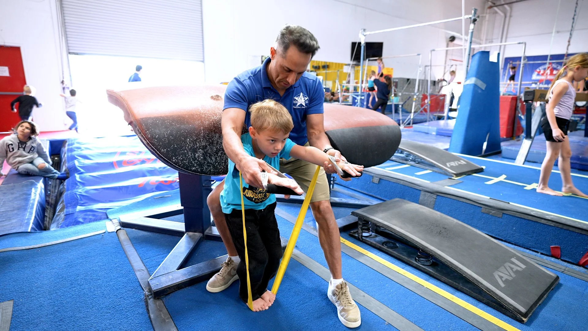 A young boy is hanging upside down from the ceiling of an indoor trampoline gym, holding onto the ceiling with his legs. There are several people and gym equipment in the background.