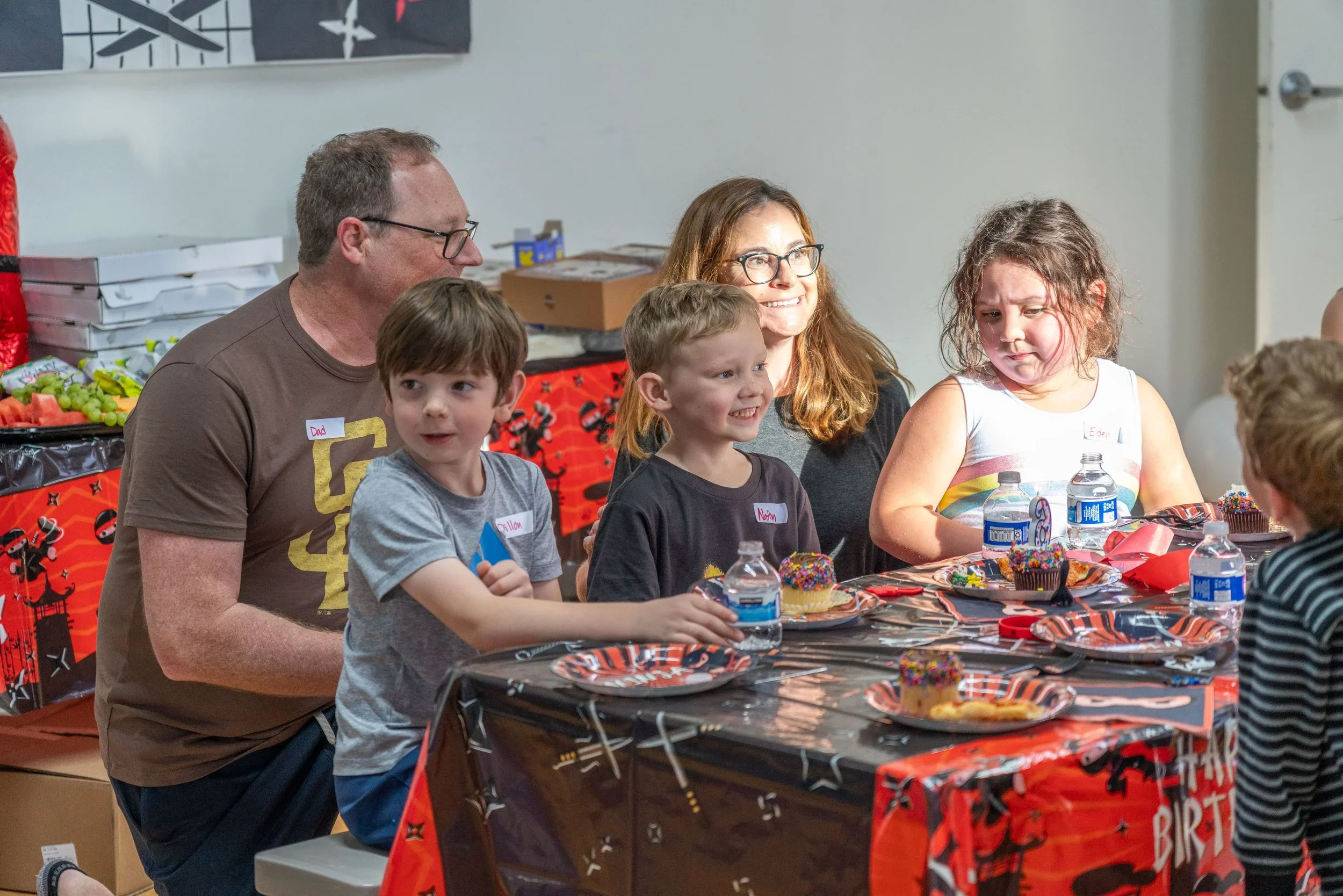A group of people, including children and adults, standing together on a blue gymnasium floor. They are smiling and wearing blue shirts or jackets, with a banner behind them reading "GYMINNY KIDS INC." and a large star. The background shows gym equipment and mirrors.