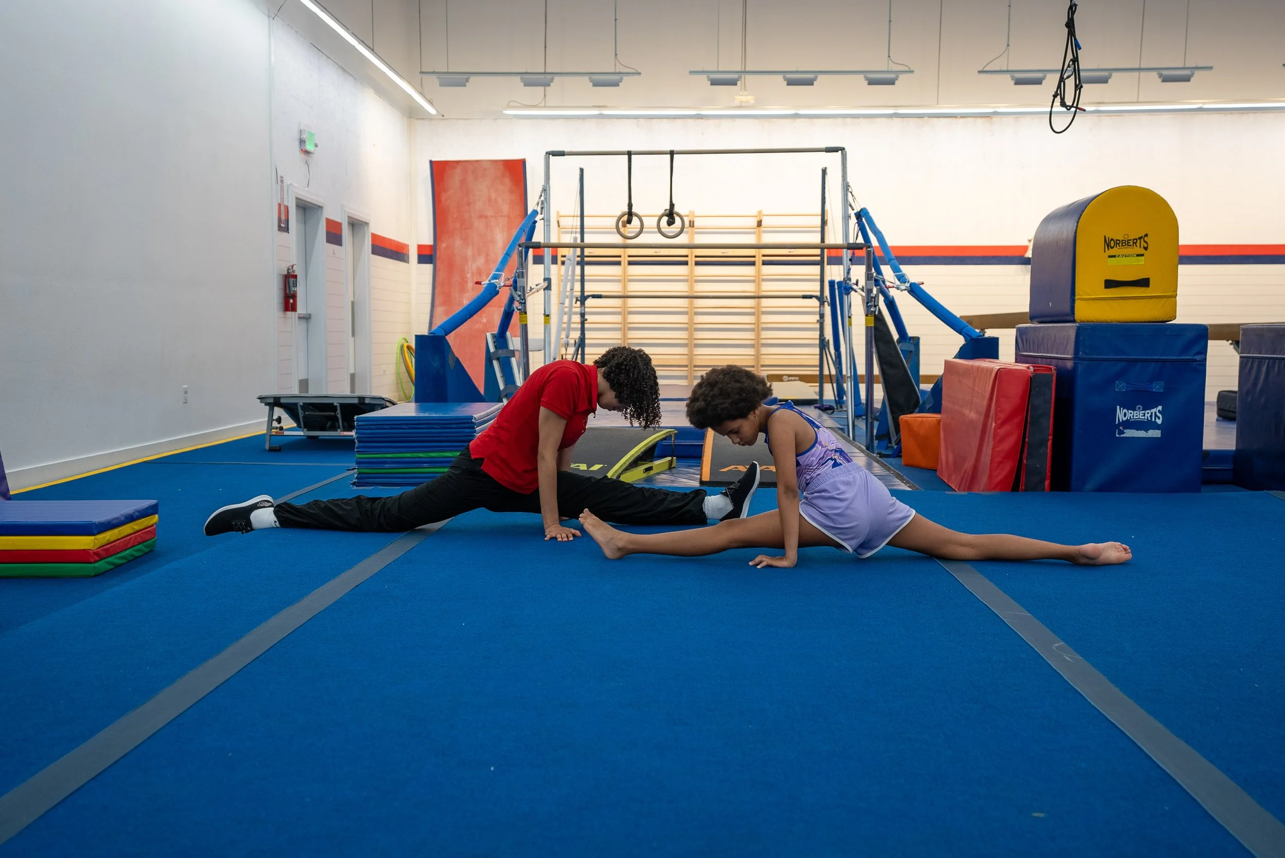 A young boy is hanging upside down from the ceiling of an indoor trampoline gym, holding onto the ceiling with his legs. There are several people and gym equipment in the background.