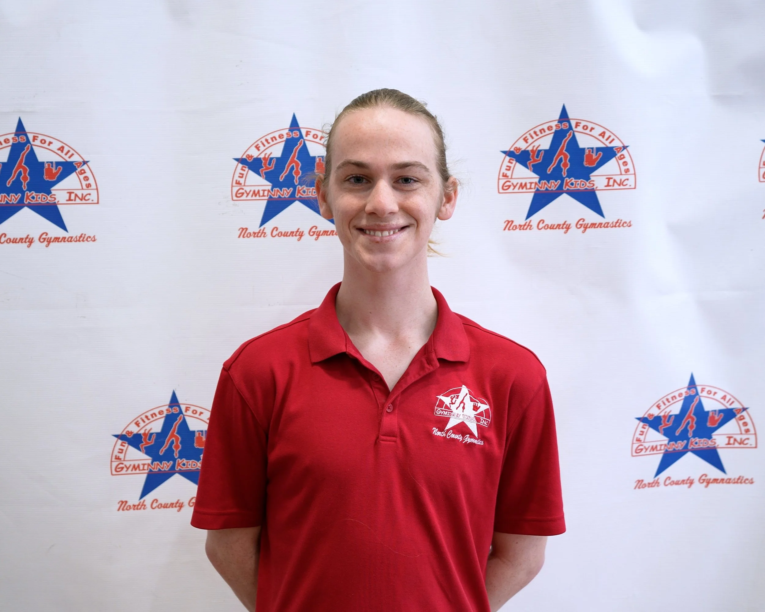 Young female gymnast standing in front of a backdrop with Gyminy Kids Inc. logo, wearing a red polo shirt with the same logo, smiling at the camera.