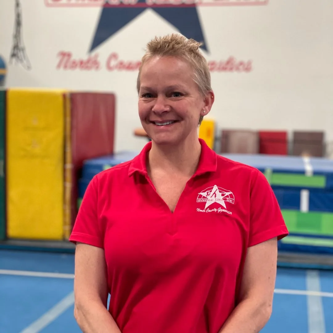 Woman with short blonde hair smiling, wearing a red polo shirt with a gymnastics logo, standing in a gymnasium with colored mats and equipment in the background.