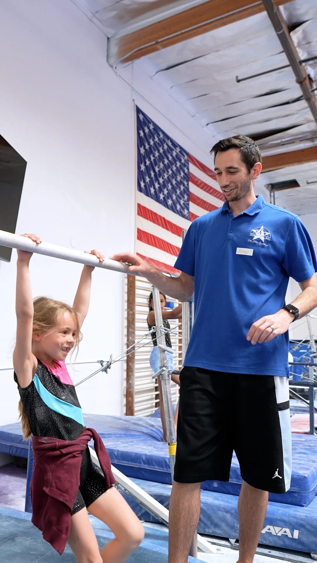 A young girl and a man in a gymnasium with an American flag in the background. The girl is hanging onto a horizontal pole while the man, wearing a blue shirt and black shorts, stands nearby, smiling.