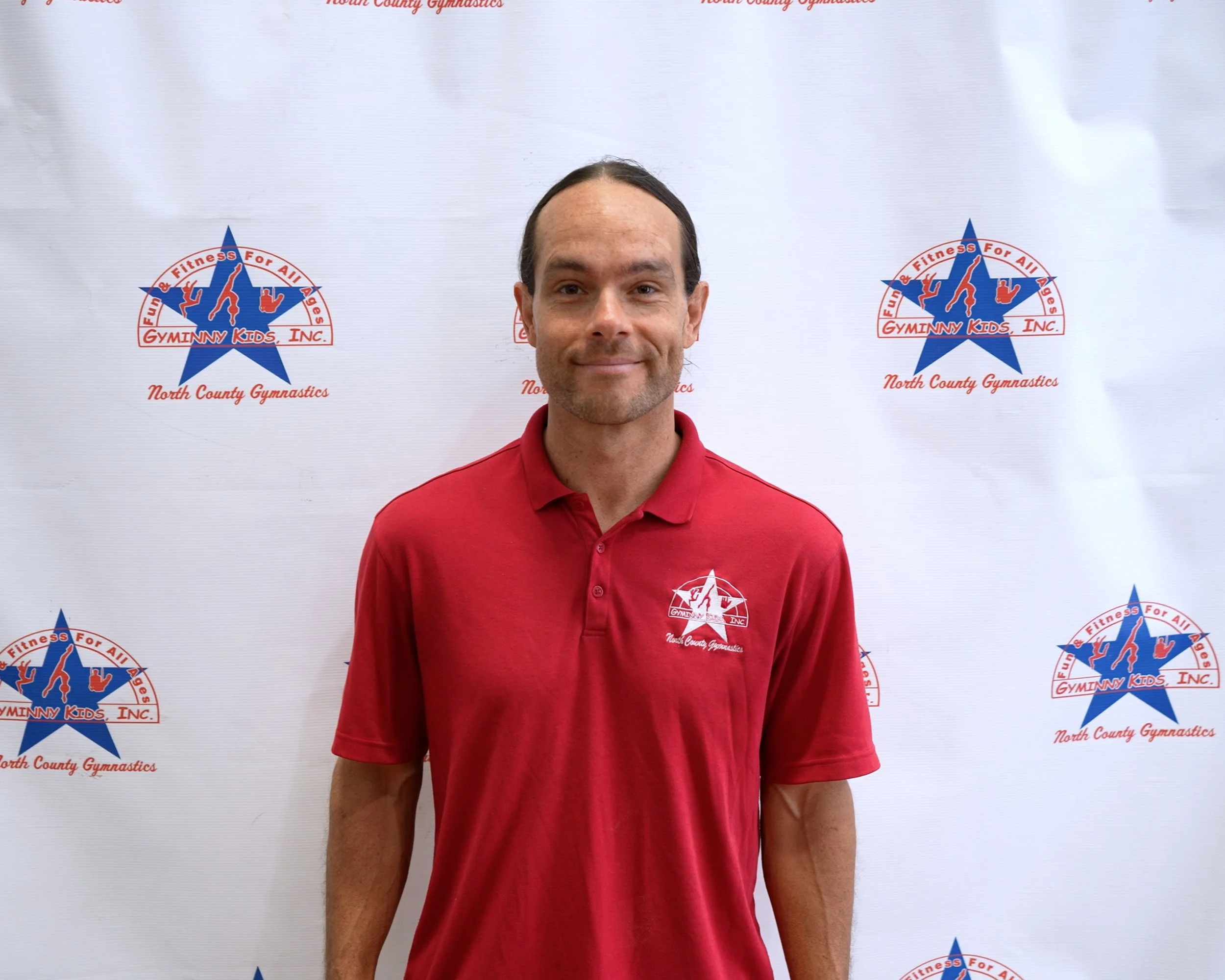 Man standing in front of a GymnNY Kids Inc. North County Gymnastics backdrop, wearing a red collared shirt with the organization’s logo.
